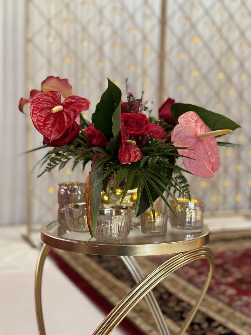 Red anthurium and rose centerpiece with palm and fern foliage on a round gold side table, ringed by mercury-glass votives against a lattice screen strung with warm fairy lights.