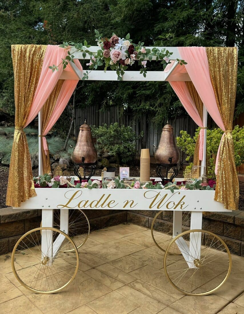 The Ladle n Wok chai cart: a white canopied cart on gold spoked wheels, draped in blush chiffon and gold sequin, topped with a rose and eucalyptus swag and set with copper samovars.