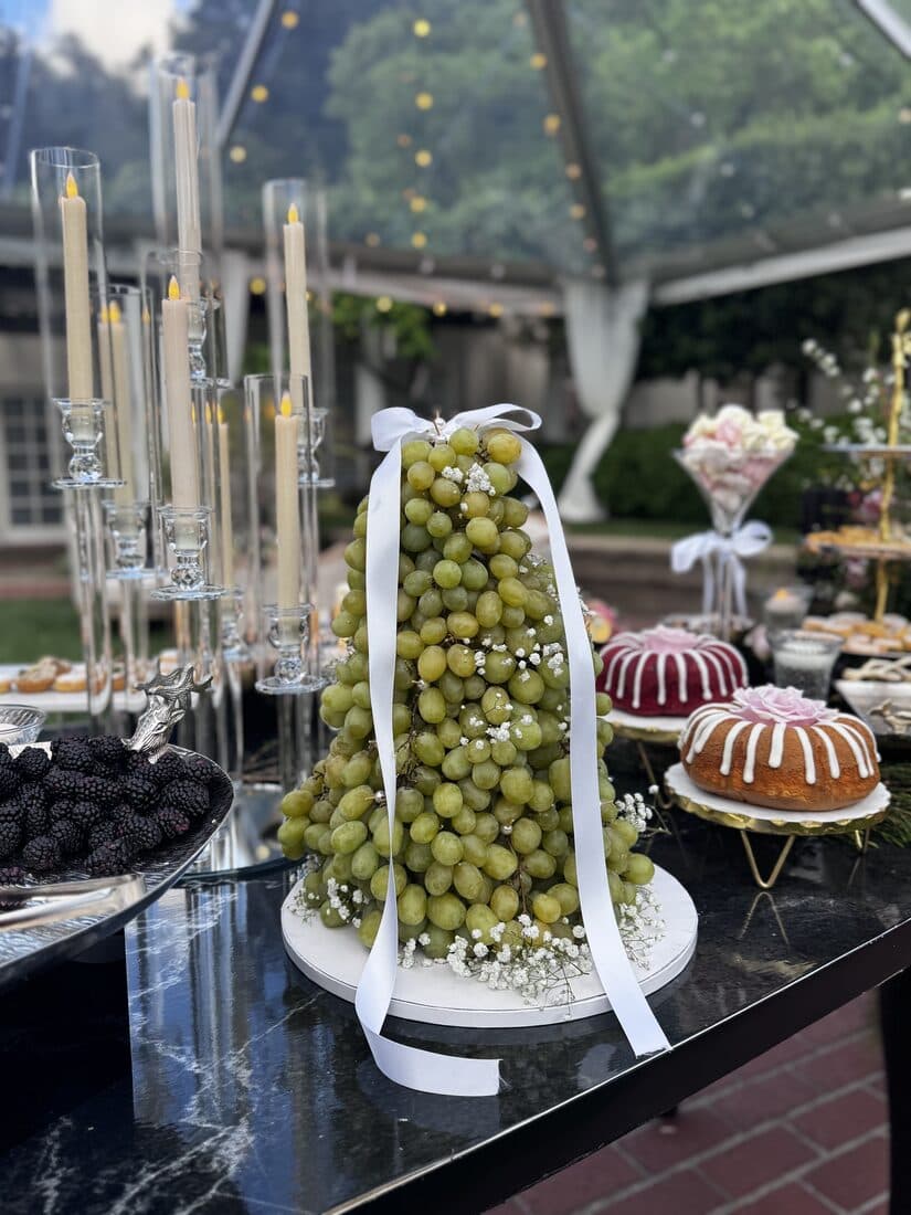 Green grape tower tied with a white ribbon on a black marble dessert table under a clear tent, flanked by tall tapered candles, glazed bundt cakes and a silver bowl of blackberries.