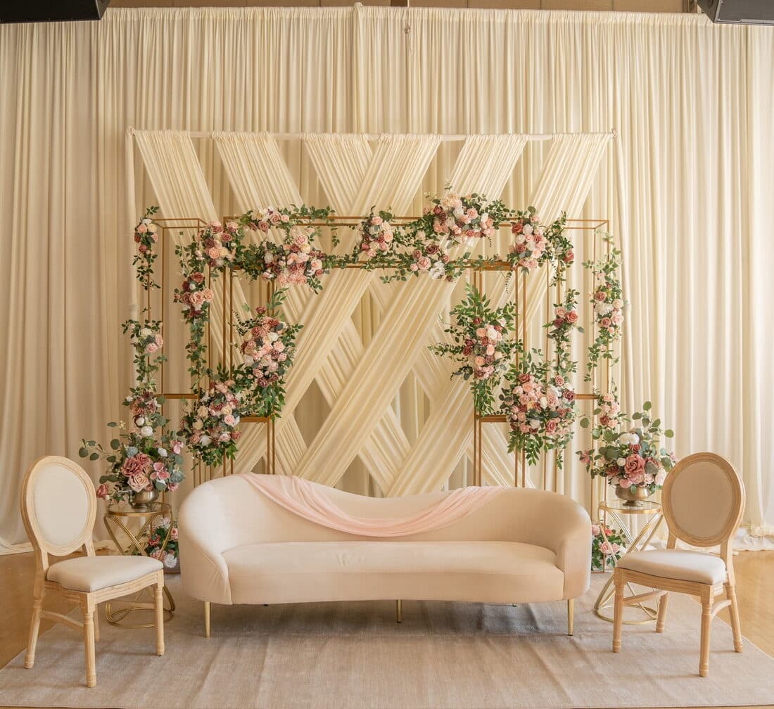 Ivory draped wedding stage with a gold frame threaded with blush, peach and cream roses and trailing greenery, set with a curved cream sofa, a blush runner and two cane-backed chairs.