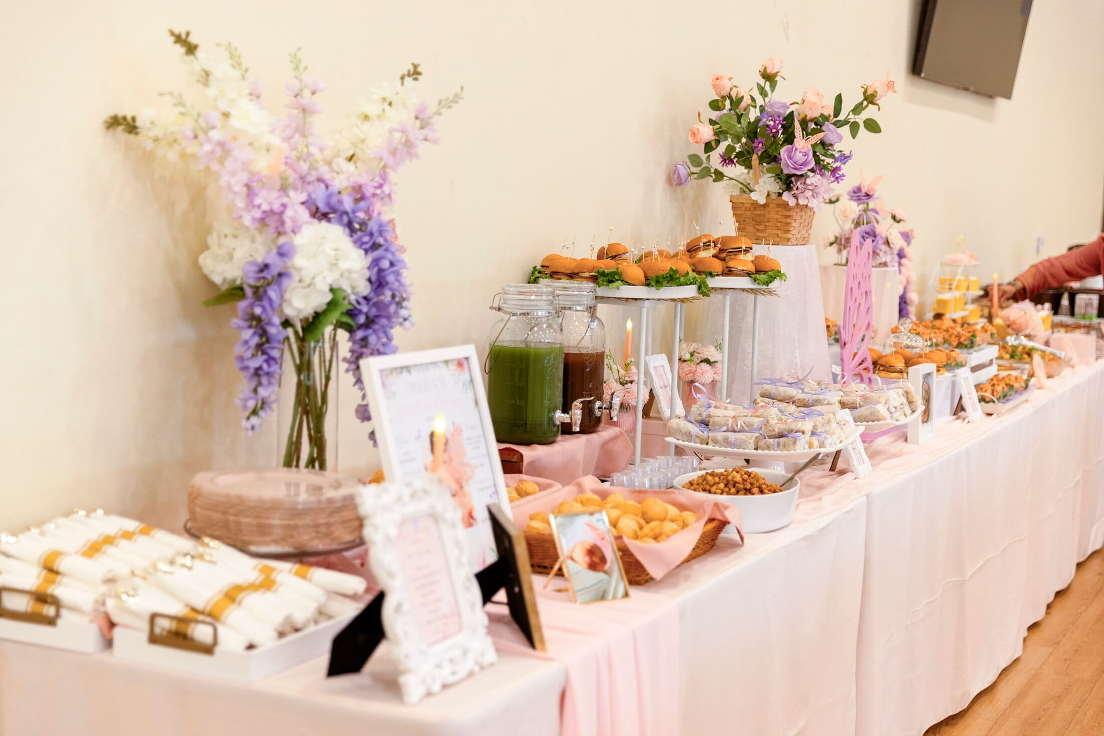 Indoor pastel dessert and savory buffet with a blush skirted table, tall purple and white floral arrangements, glass beverage dispensers of green and brown drinks, mini slider sandwiches, chickpeas, and framed menu cards.