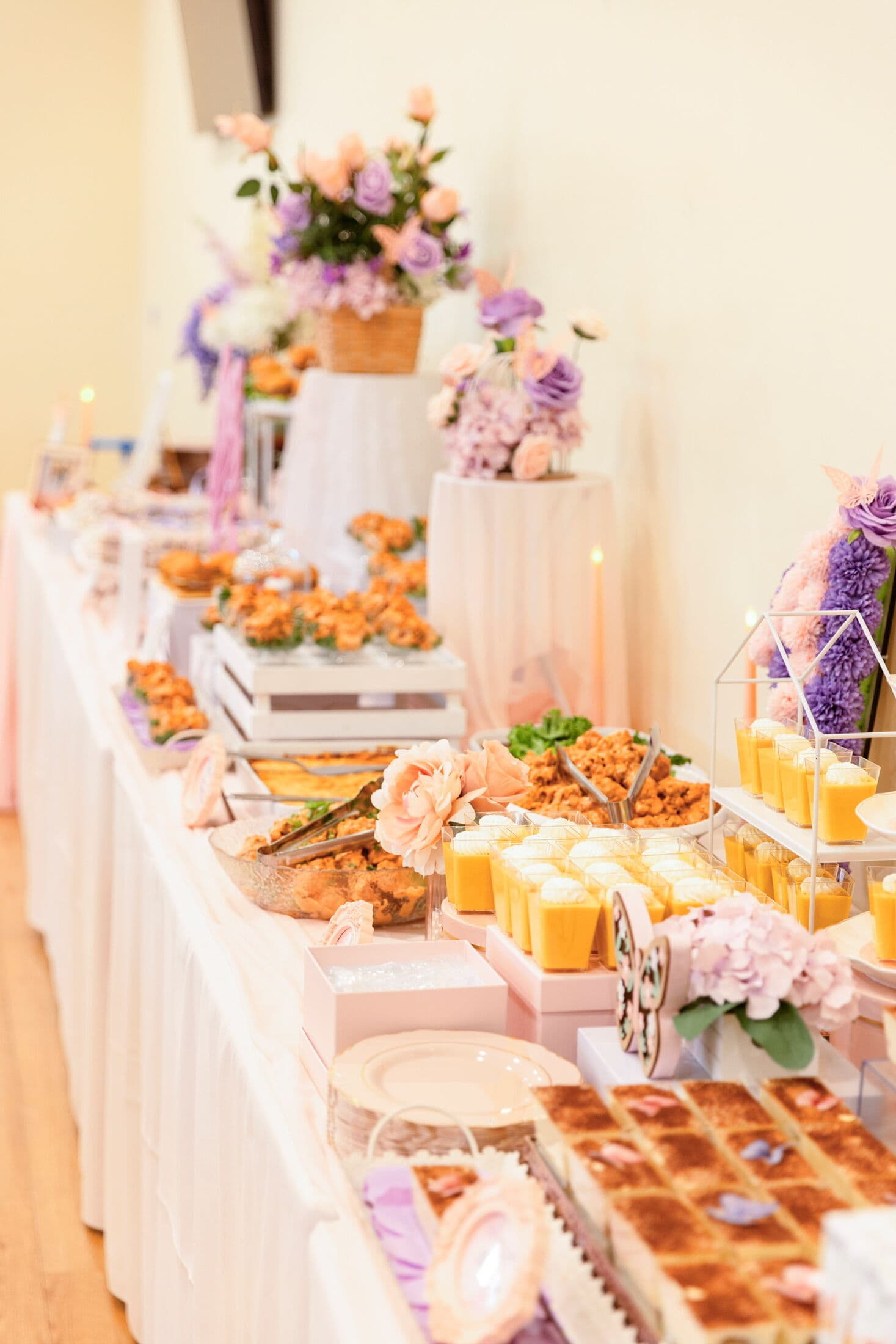 Close-up of a long blush-draped grazing table with tiered fried chicken, mango pudding cups, tres leches squares, and lavender-and-peach floral risers on white pedestals indoors.