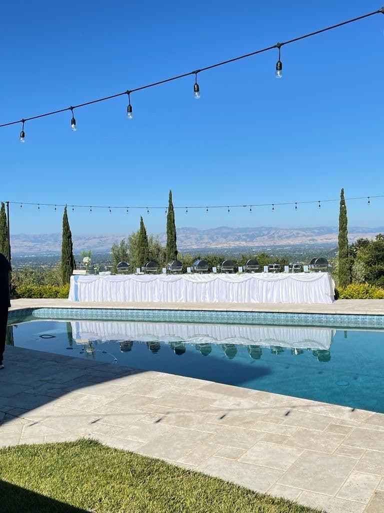 Outdoor poolside Bay Area buffet with a white ruffle-skirted table lined with silver chafing dishes, set against string lights and a hilltop view of cypress trees and distant mountains.
