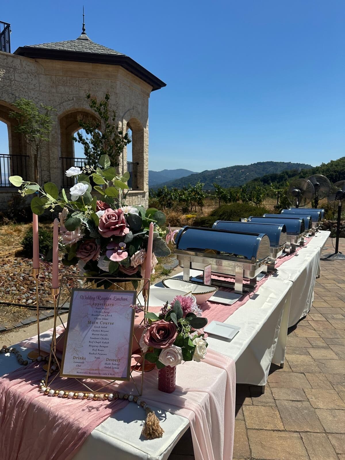 Outdoor wedding reception luncheon buffet by Ladle n Wok with a dusty-rose runner, navy chafing dishes, mauve rose centerpieces, taper candles, and a printed menu framed beside a stone villa and vineyard hills.