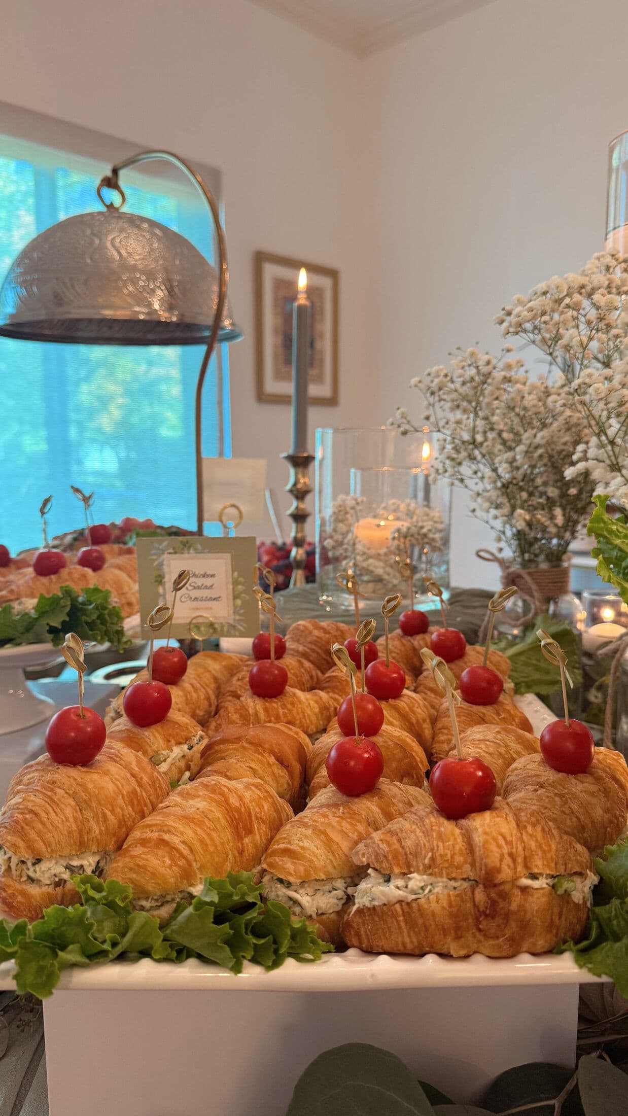 Platter of chicken salad croissant sandwiches topped with cherry tomatoes on decorative picks over leafy lettuce, with a labeled card, baby's breath, and a pillar candle in a bright indoor setting.