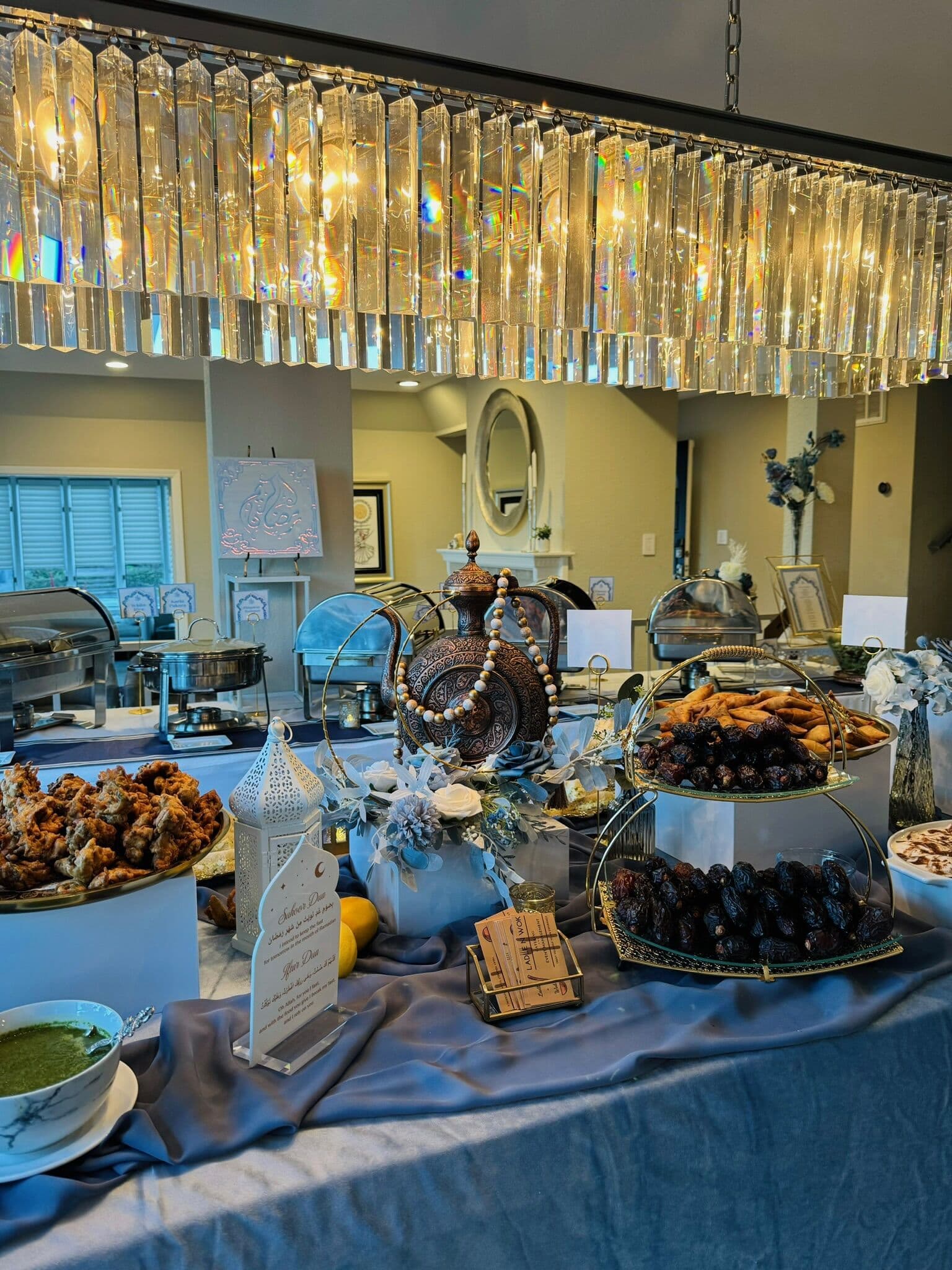 Indoor buffet beneath a crystal chandelier with a dusty-blue linen, silver chafing dishes, a footed tray of dates, fried appetizers, and an ornate silver serving dome.