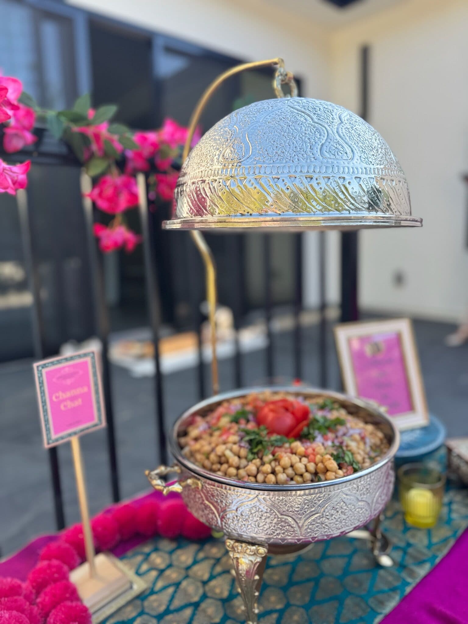 Ornate silver chafing dish with a raised etched dome revealing chana chaat garnished with tomato and herbs, beside a pink Channa Chaat sign and fuchsia bougainvillea at an outdoor Bay Area mehndi.