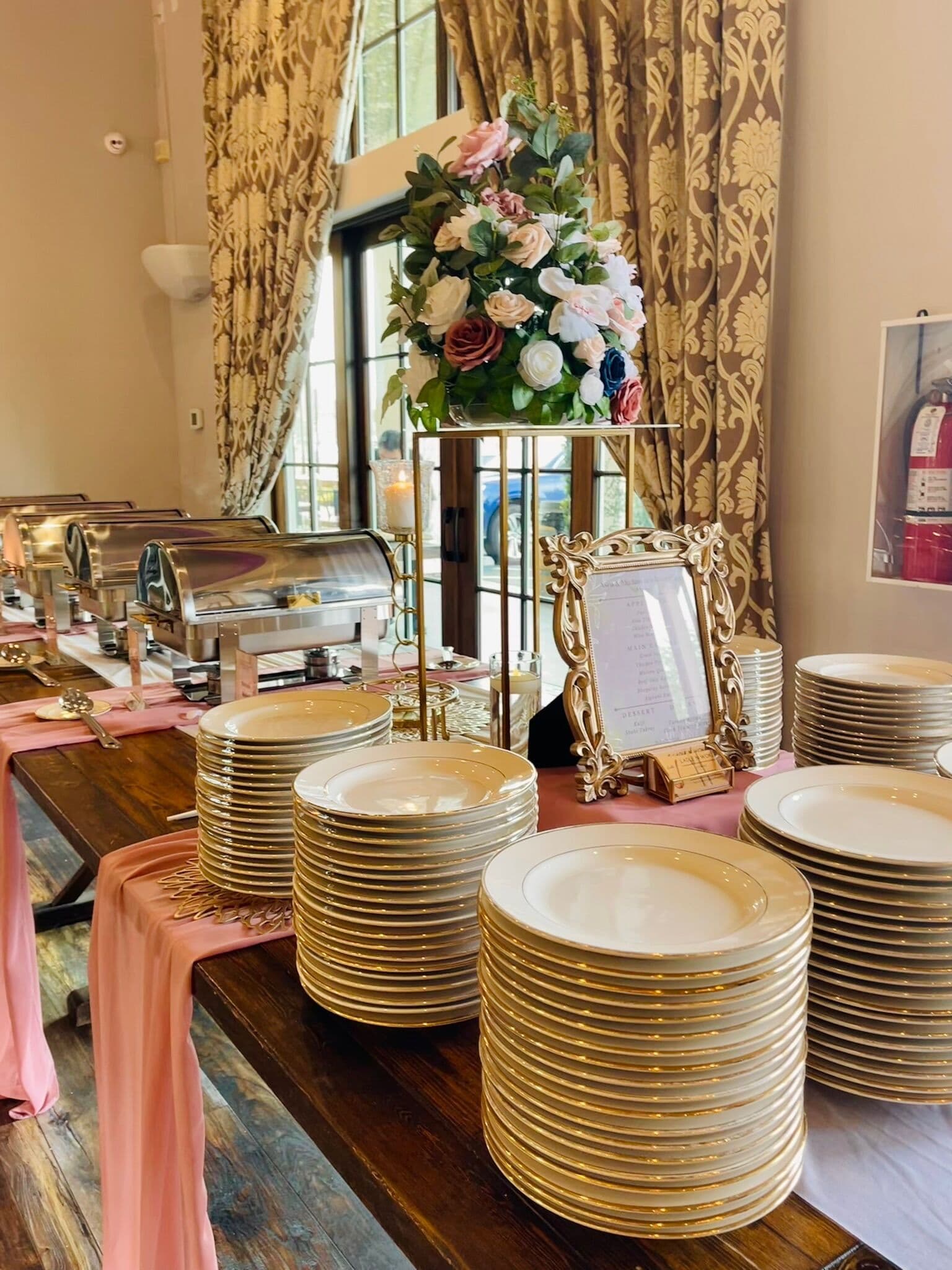 Rustic wood buffet table with tall stacks of gold-rimmed china plates, silver chafing dishes, a blush runner, a framed menu, and a rose-and-hydrangea arrangement against damask draperies.