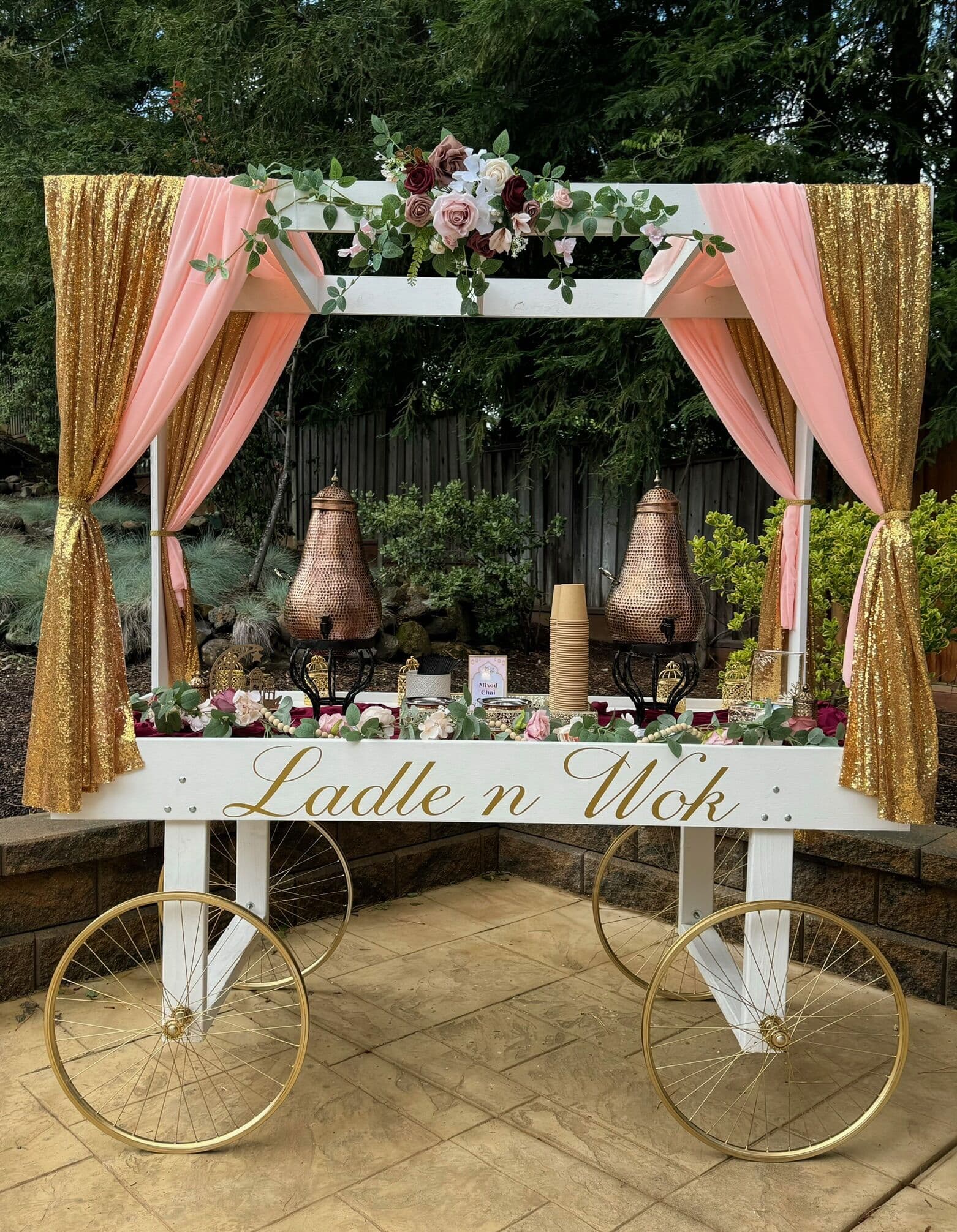 White and gold carriage-style dessert cart lettered "Ladle n Wok" with blush and gold drapery, a rose-and-eucalyptus swag, copper serving vessels, and spoked wheels set on a backyard patio.