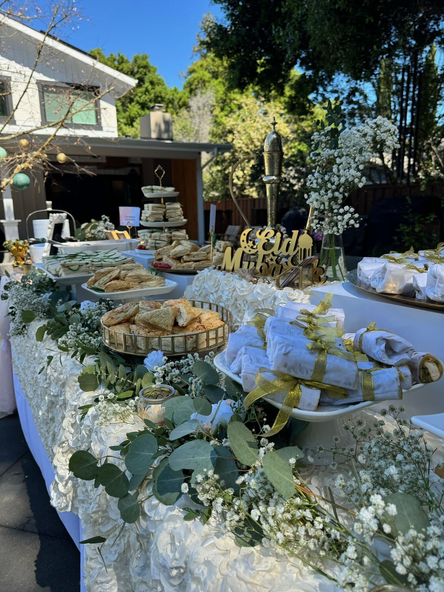 Outdoor Eid Mubarak grazing table dressed in ivory textured linen and eucalyptus with baby's breath, tiered savory pastries, scones, and gold-ribbon-tied napkin bundles in a Bay Area backyard.