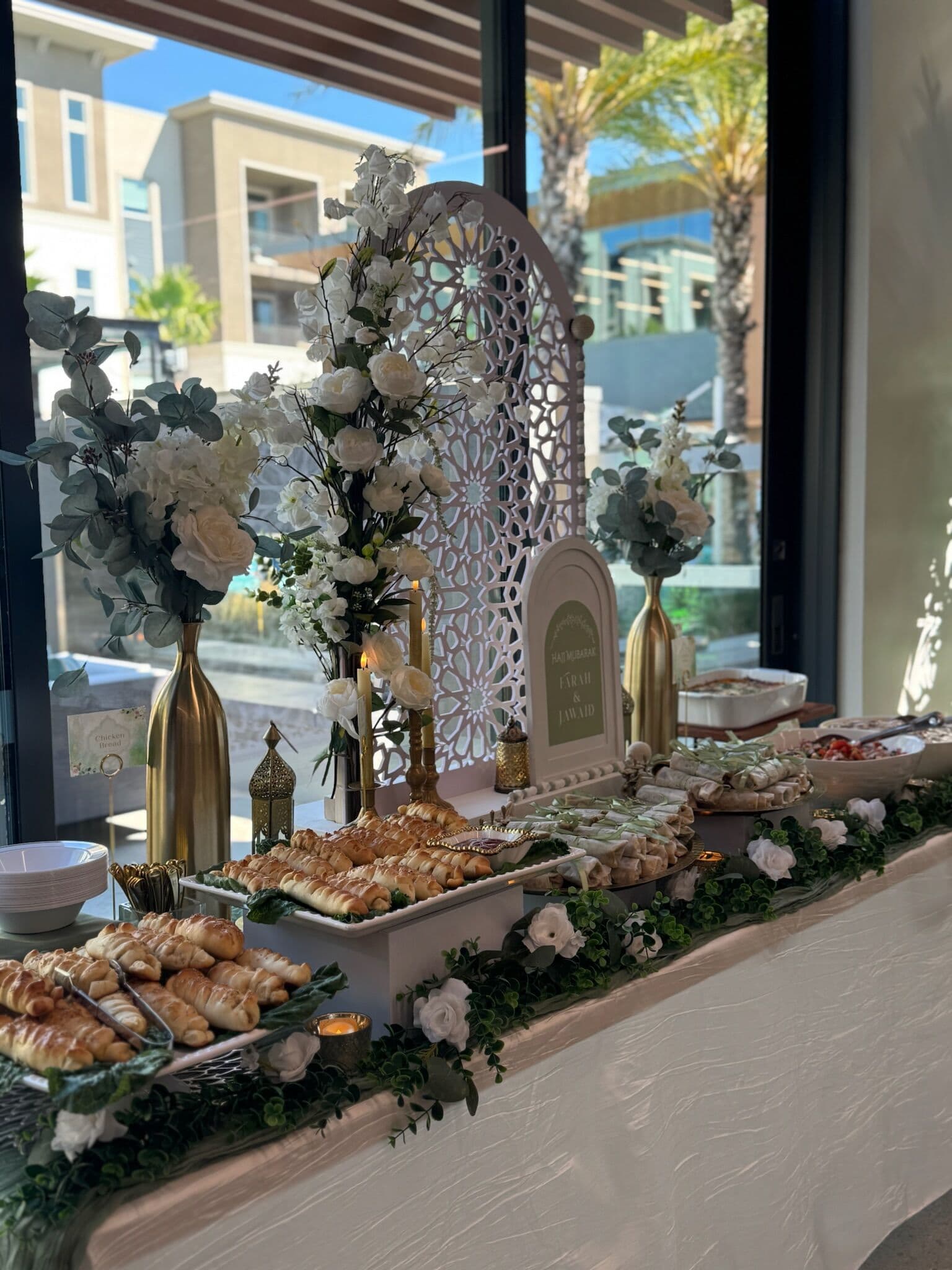 Elegant Hi-Tea buffet against floor-to-ceiling windows with a white Moroccan-arch backdrop, gold vases of white roses and eucalyptus, croissants, and rolled savory pastries on a greenery-lined table.