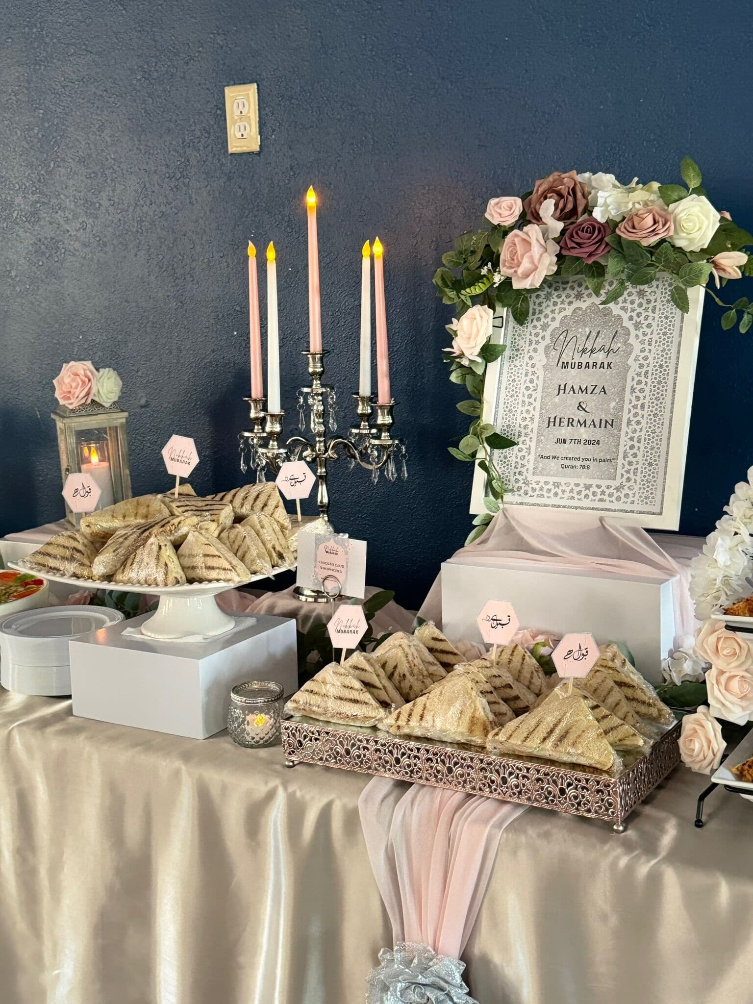 Nikah Mubarak dessert table for Hamza and Hermain against a navy wall, with a silver candelabra, blush taper candles, mauve roses, a framed sign, and tiered platters of samosas and folded pastries.