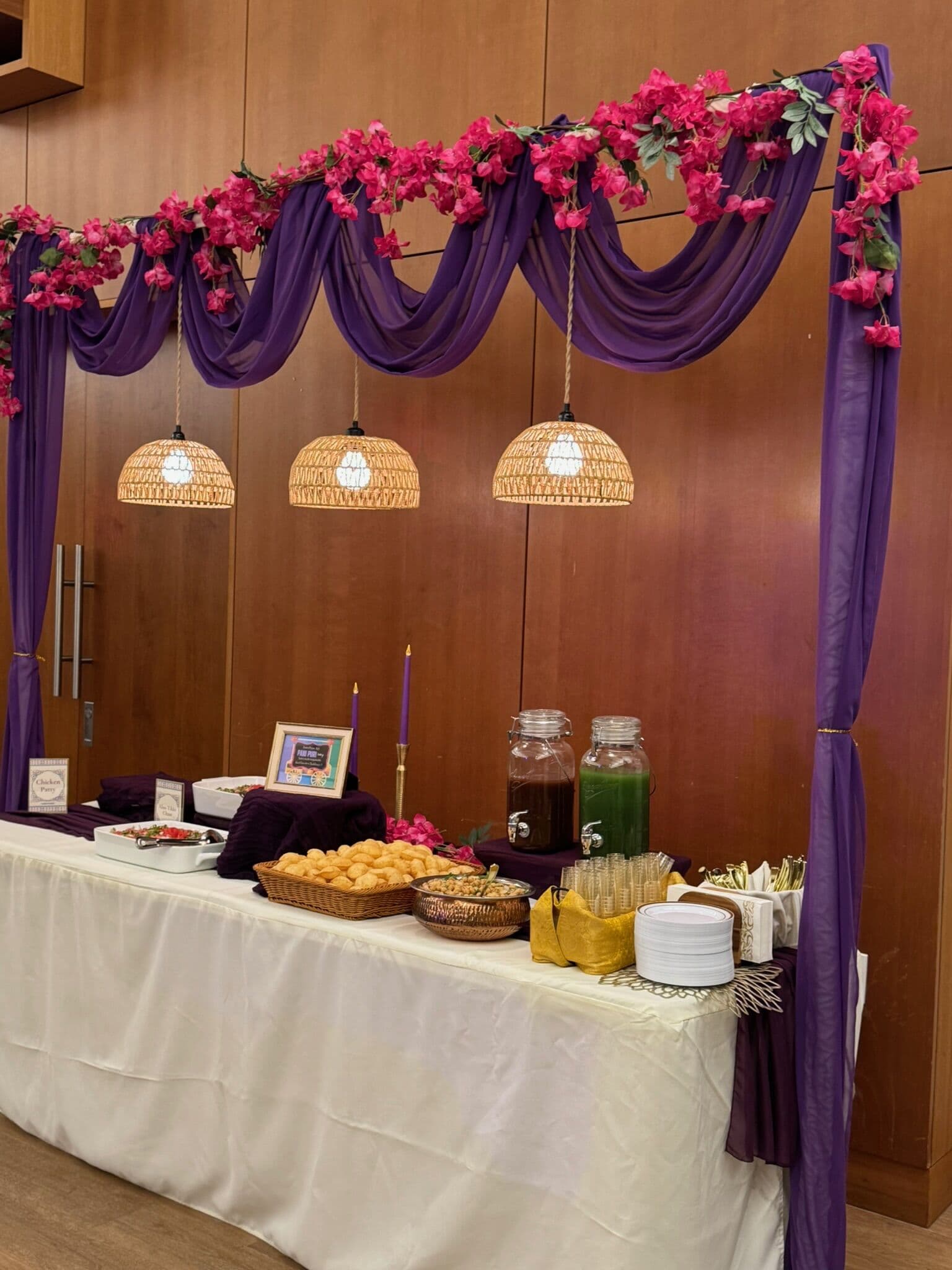 Mehndi pani puri and chaat station under a purple drape and fuchsia bougainvillea garland with woven pendant lamps, jars of tamarind and mint chutney, a basket of puri shells, and stacked plates on a white-skirted table.