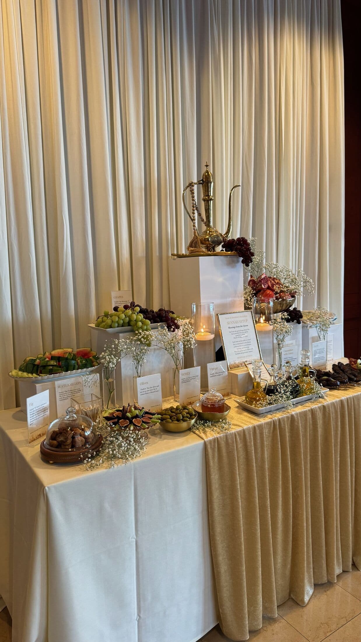 Sunnah-inspired grazing table on white pedestals with a brass Arabic dahleh pitcher and prayer beads, arranged watermelon, grapes, figs, olives, dates, honey, and olive oil with baby's breath against ivory drapery.