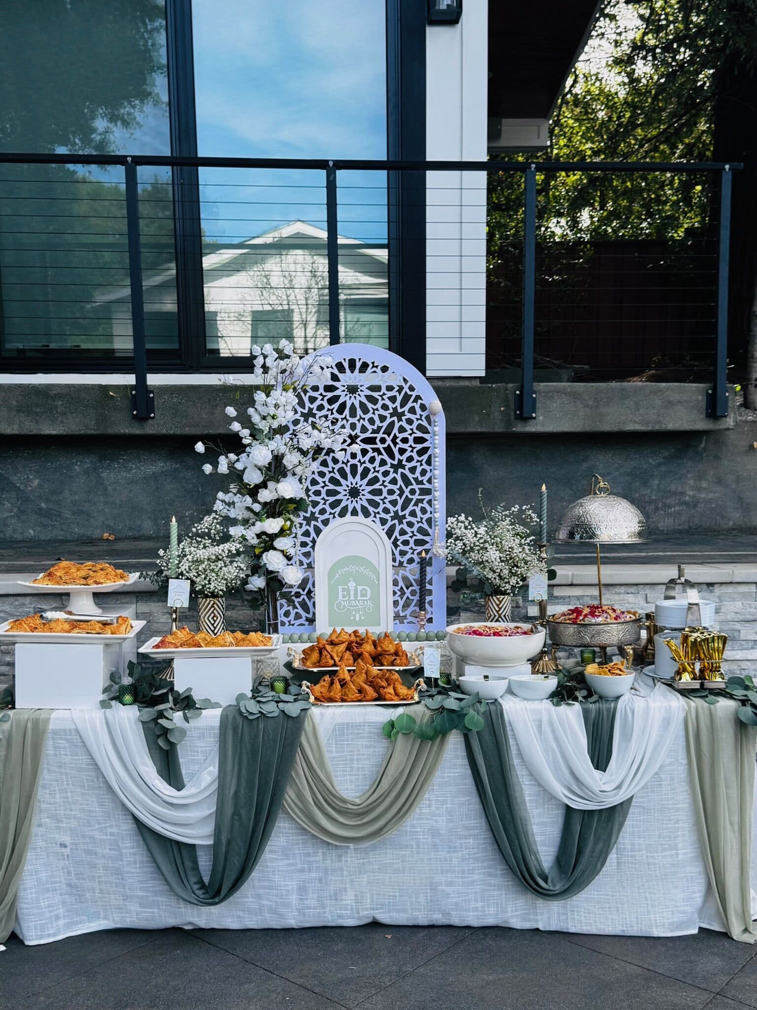 Outdoor Eid buffet with a white Moroccan-arch sign, sage-green draped linen, baby's breath and white florals, platters of fried pakoras and samosas, chaat, and a silver serving dome by a modern home.