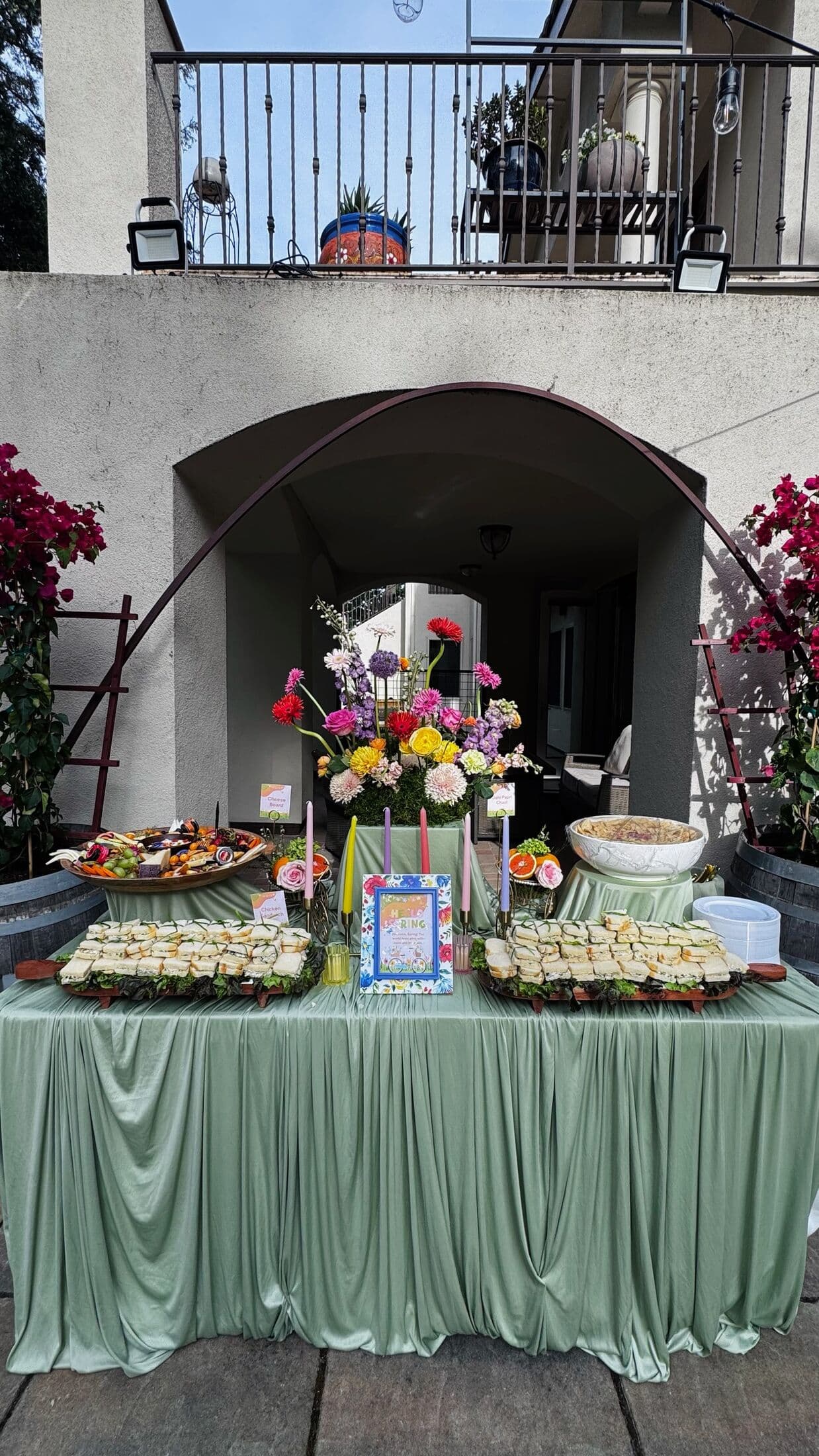 Outdoor sage-green skirted buffet under a stucco archway with a vivid pink, yellow, and orange floral centerpiece, tea sandwiches, a charcuterie platter, and a footed fruit bowl in a Bay Area courtyard.