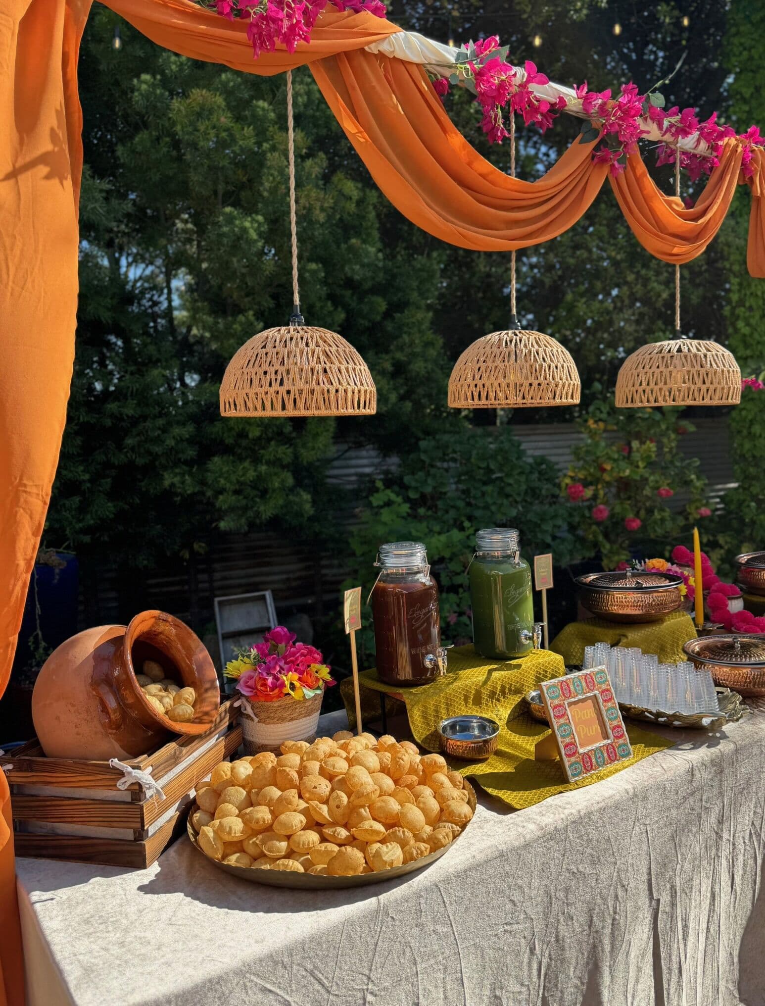 Outdoor evening chaat station with orange drapery and fuchsia bougainvillea, woven pendant lamps, jars of tamarind and mint chutney, a clay pot, and a platter of golgappa shells on a natural linen table.