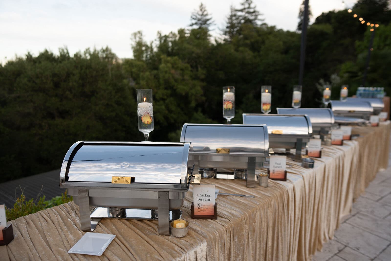 Row of gleaming silver roll-top chafing dishes on a champagne velvet-draped table with floating candles and menu cards for Chicken Biryani and Veal Karahi, set outdoors against a wooded Bay Area hillside at dusk.