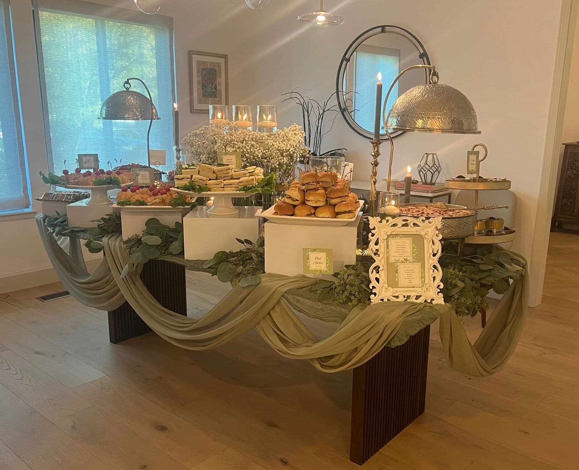 Indoor buffet on a dark wood table with sage-green draped swags, white pedestals of tea sandwiches and slider rolls, a raspberry-topped platter, baby's breath, taper candles, and silver serving domes near a round mirror.