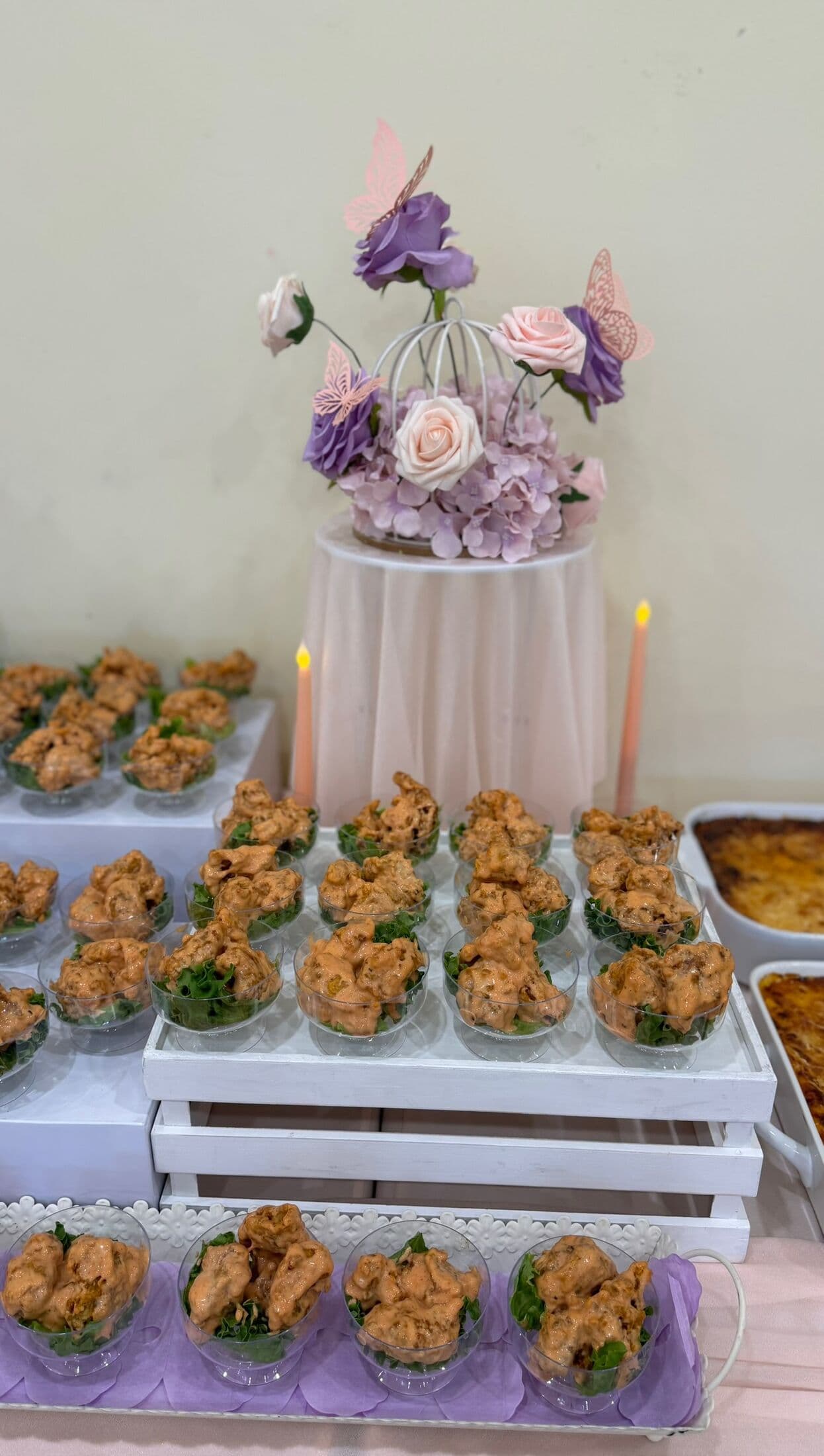 A tiered white-crate display of halal fried chicken pakoras plated in individual clear cups over fresh greens, crowned with a floral centerpiece of purple and blush roses and paper butterflies.