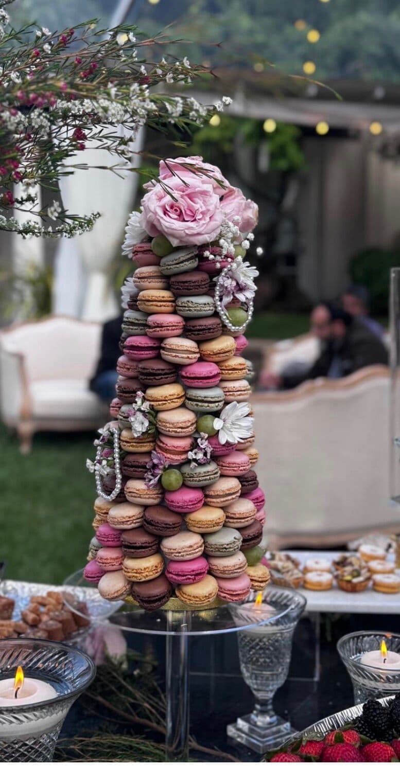 A tall macaron tower in ombre shades of pink, brown and cream, topped with pink roses and draped with pearl strands, set on a garden dessert table alongside dishes of fudge and berries under string lights.