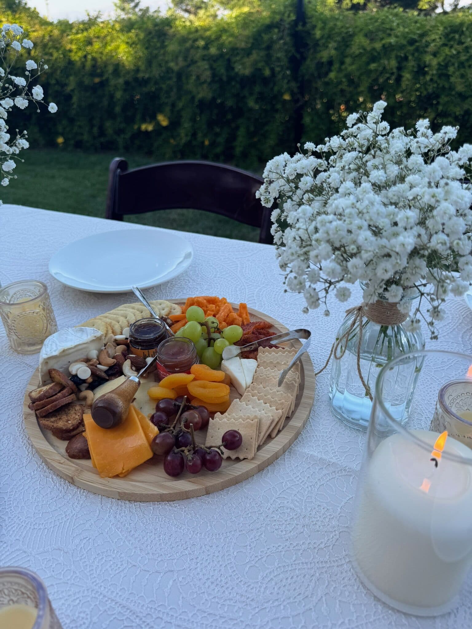 A round wooden grazing board arranged with sliced cheeses, crackers, dried apricots, grapes, nuts and honey, styled on a white linen outdoor table with a baby's breath bouquet and a lit candle in the Bay Area.