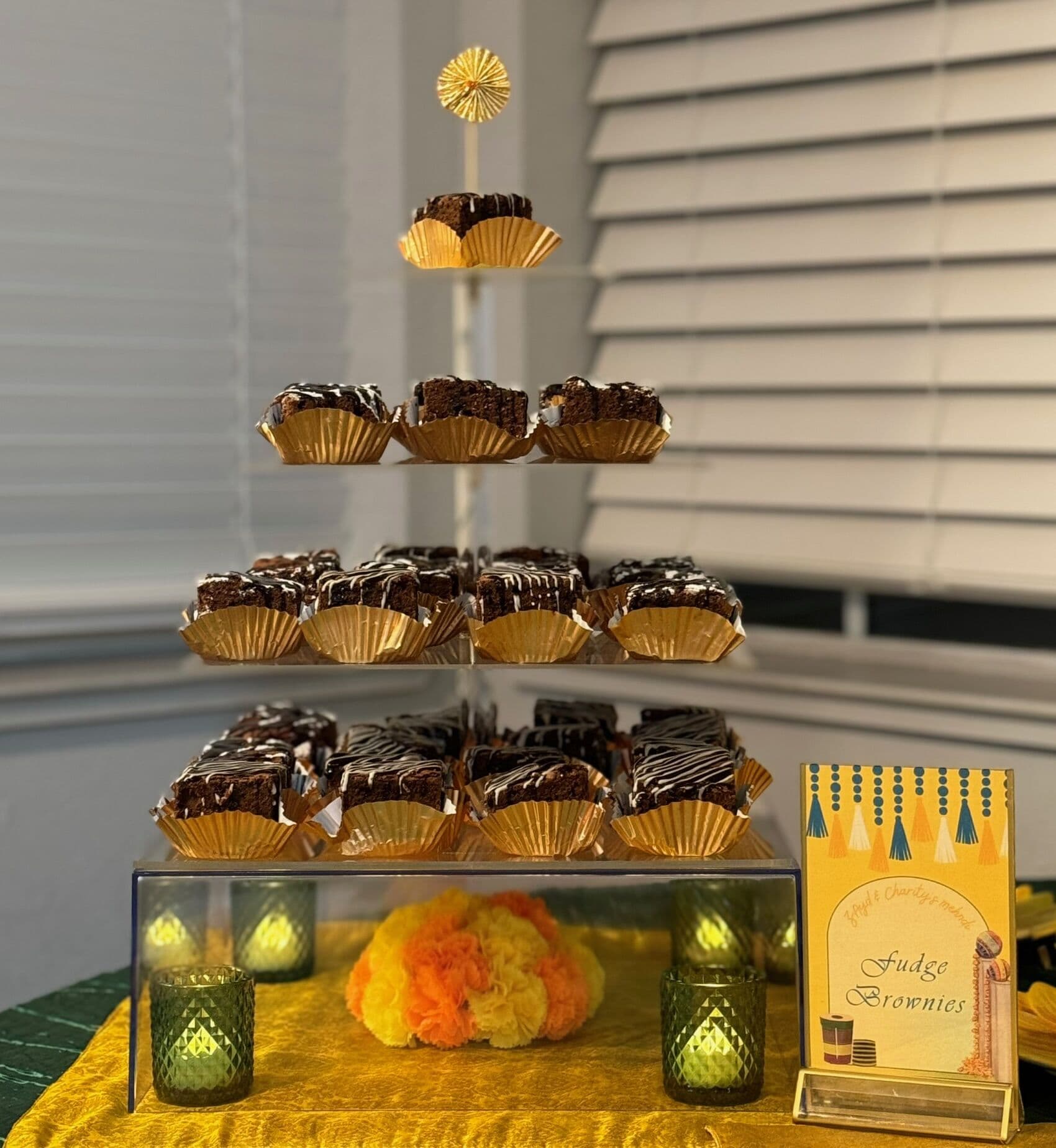 A four-tier acrylic dessert stand displaying chocolate-drizzled fudge brownies in gold foil cups, with a Fudge Brownies menu card, orange pom-poms and green votive candles on a gold cloth.