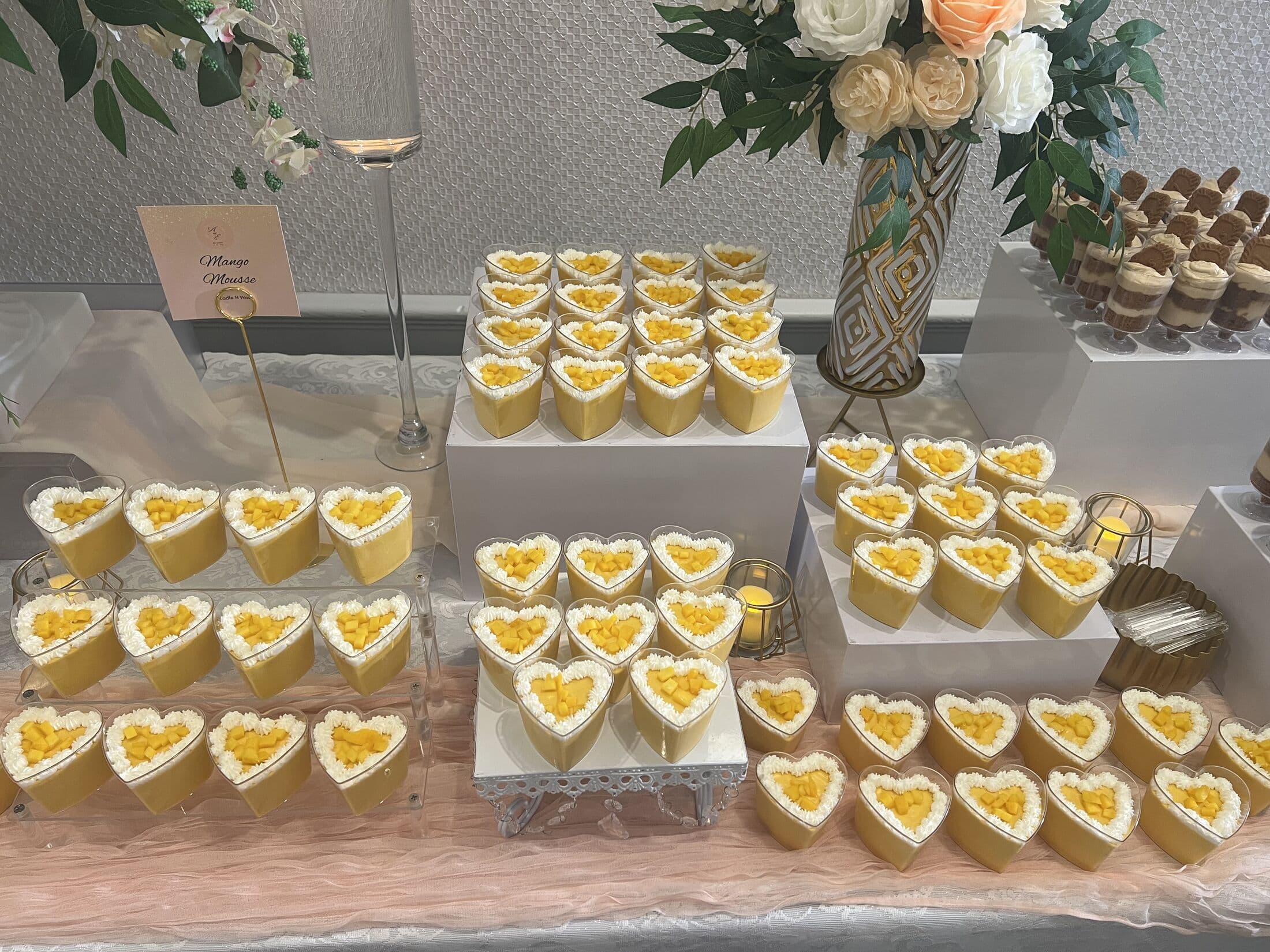 A dessert table of heart-shaped cups of mango mousse topped with cream and diced mango, arranged in rows on tiered white risers with a menu card and a floral arrangement of white and peach roses.