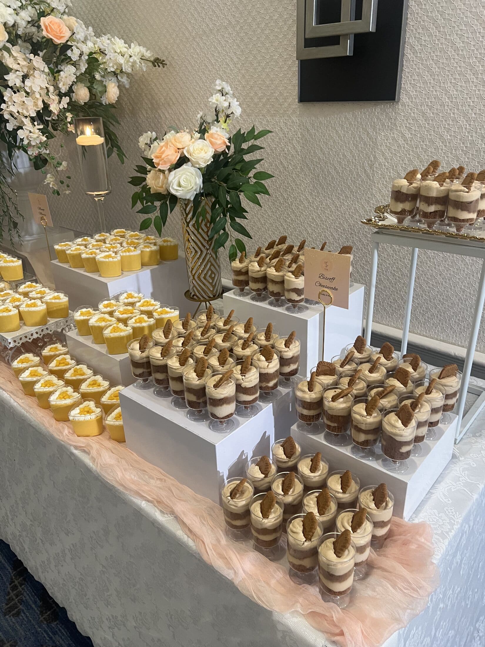 A layered dessert display combining heart-shaped mango mousse cups and tall clear cups of tiramisu-style cream dessert on white risers, styled with peach and white rose bouquets and a menu placard.