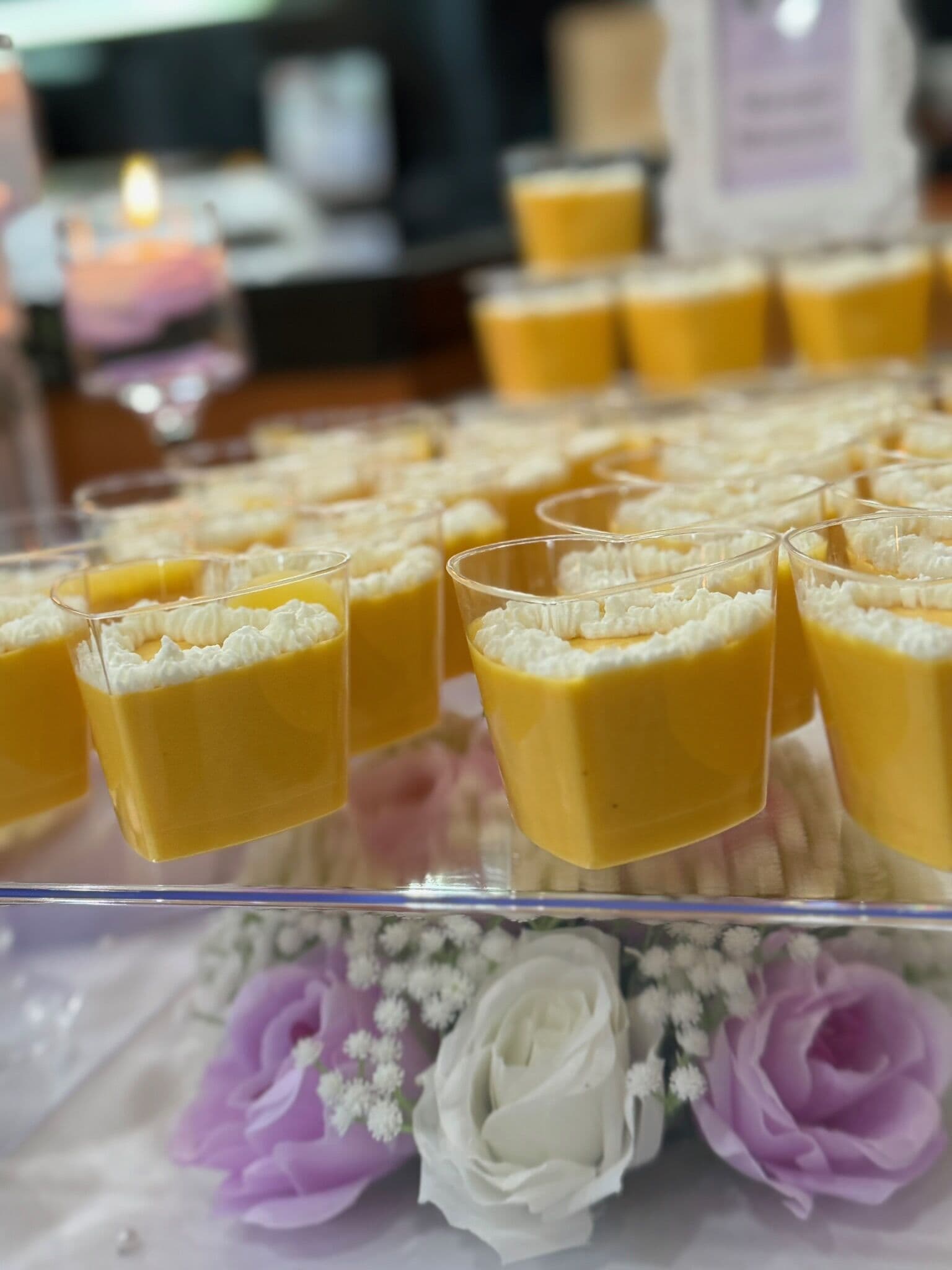 A close view of individual mango mousse shot cups piped with white cream, lined up on a clear tray decorated with purple and white roses and baby's breath at a catered event.