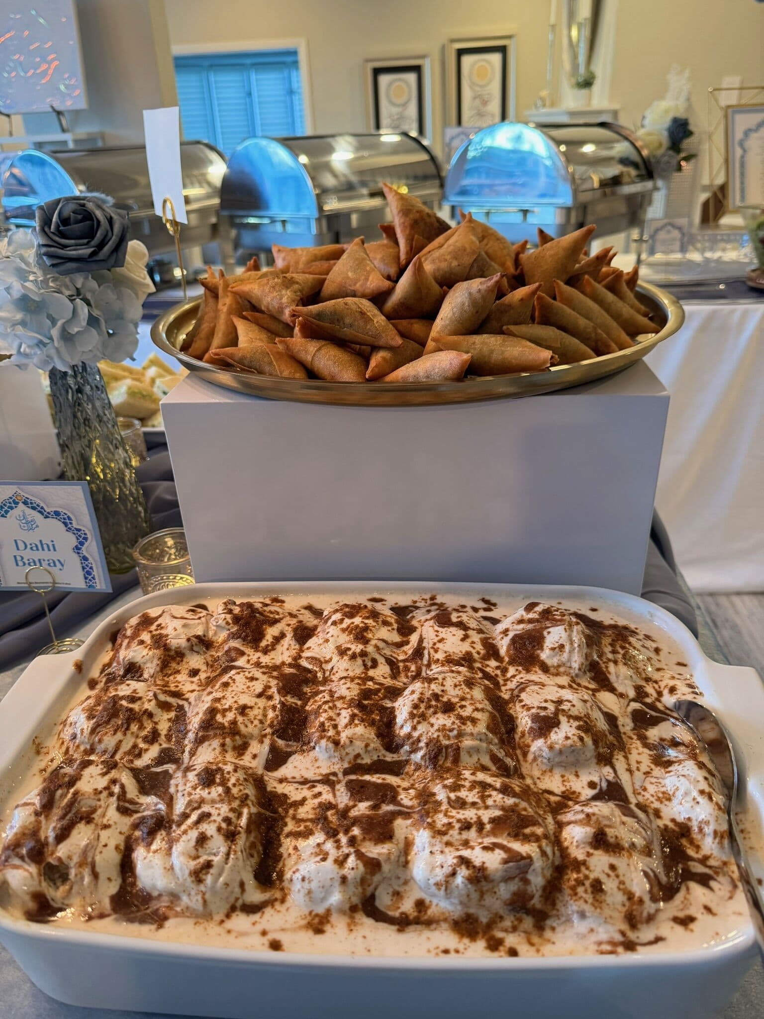 A catered dessert and appetizer setup with a white dish of dahi baray topped with cinnamon-dusted yogurt in the foreground and a gold tray of fried samosas on a riser behind, chafing dishes lining the back.