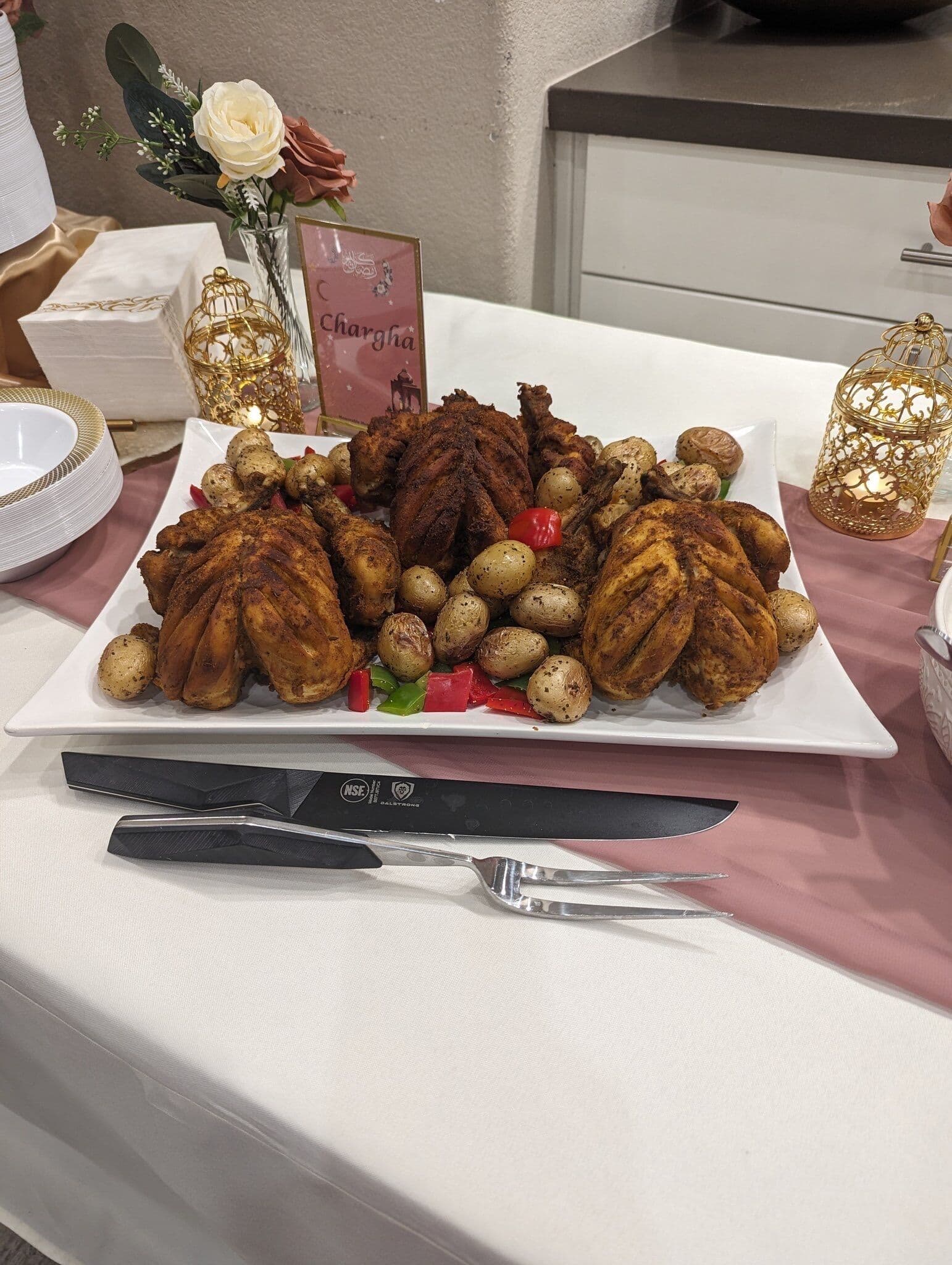 A whole roasted chicken chargha plated on a square white platter with baby potatoes and bell peppers, presented with a carving knife and fork beside a Chargha place card and gold lanterns.