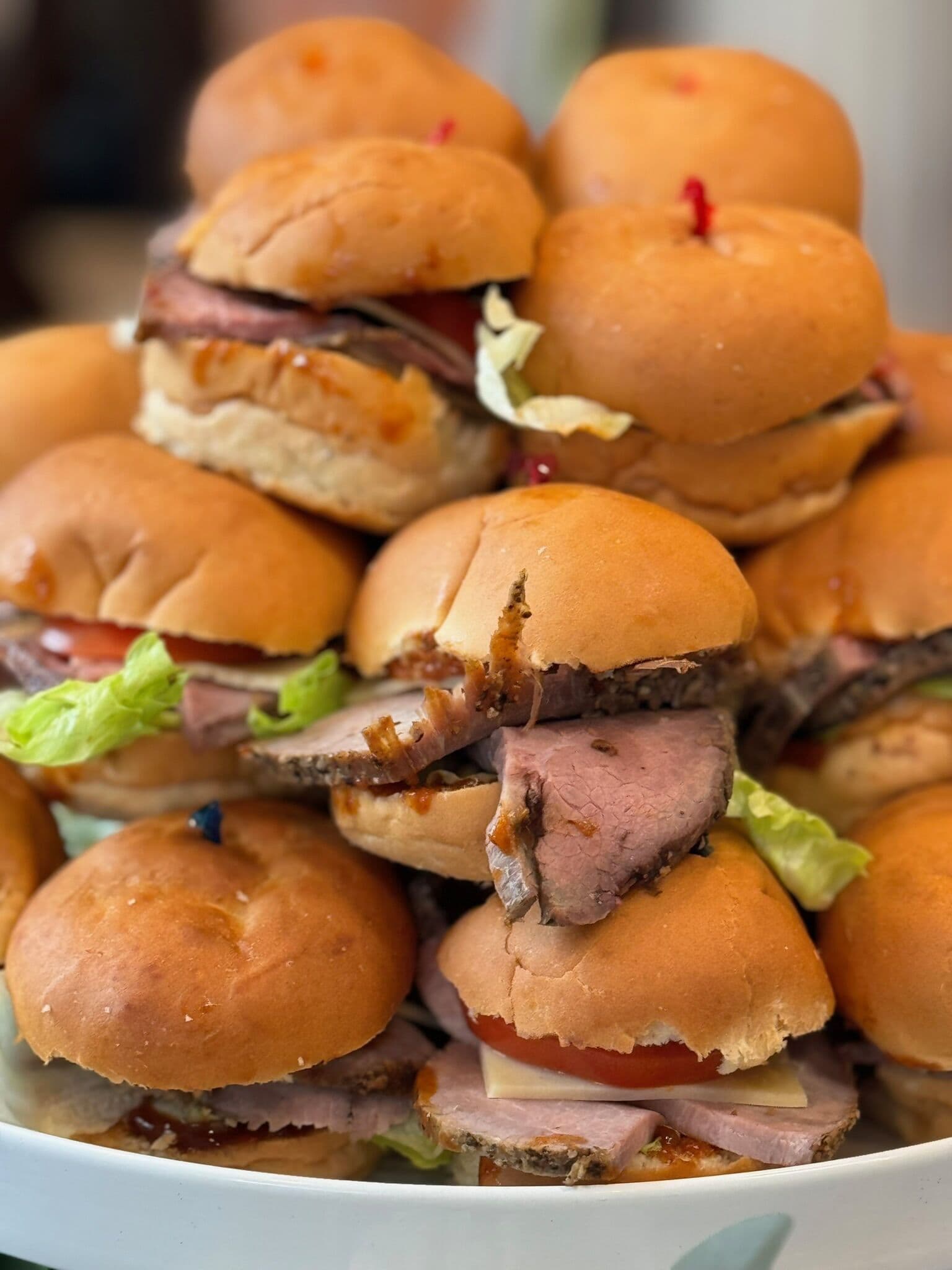 A tall stack of sliced roast beef sliders on soft dinner rolls with lettuce and tomato, secured with cocktail picks and mounded on a white plate.