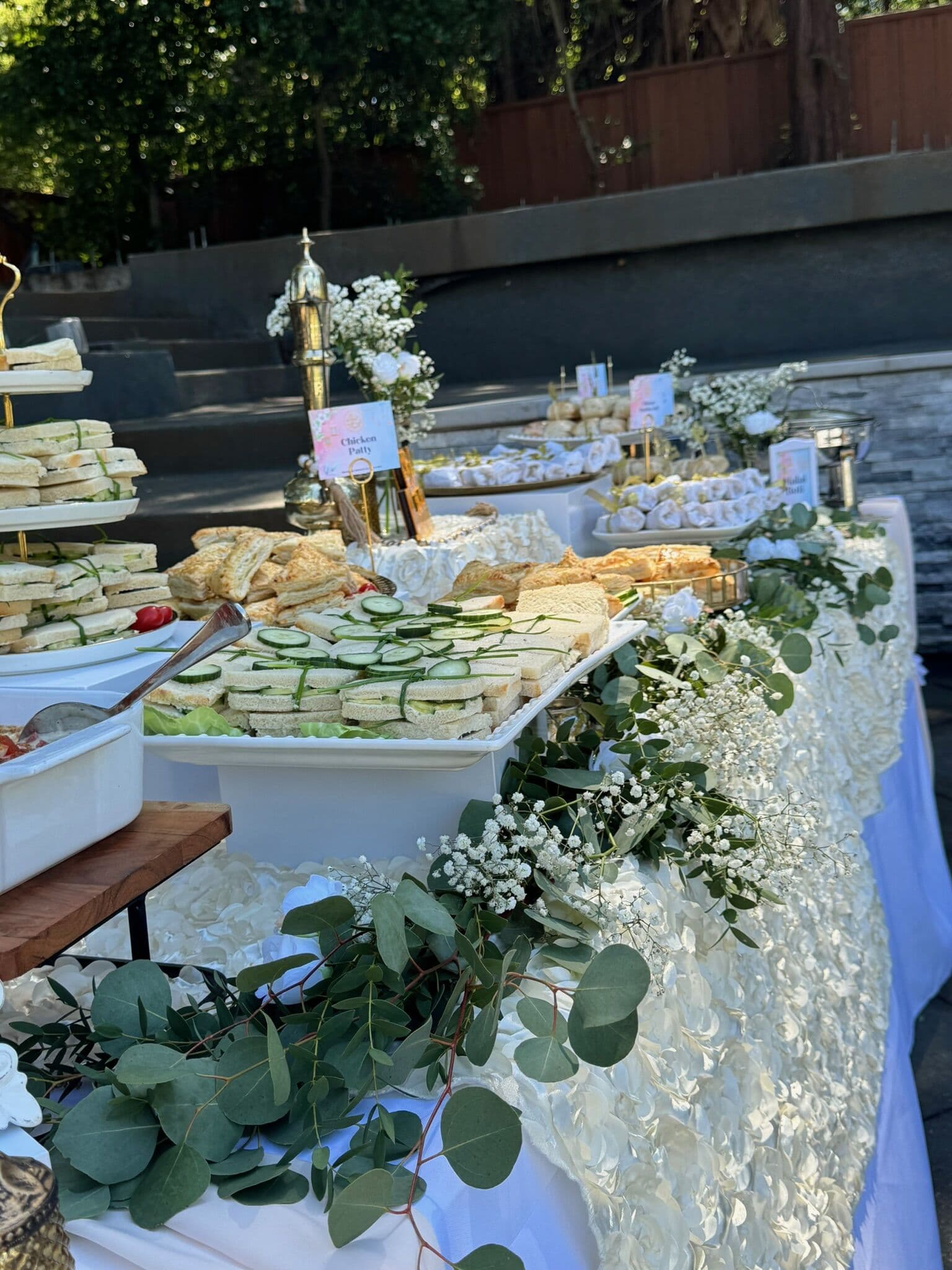 A long outdoor Hi-Tea buffet lined with platters of finger sandwiches and pastries, menu cards and lush eucalyptus and baby's breath garlands running along a white draped table in the Bay Area.