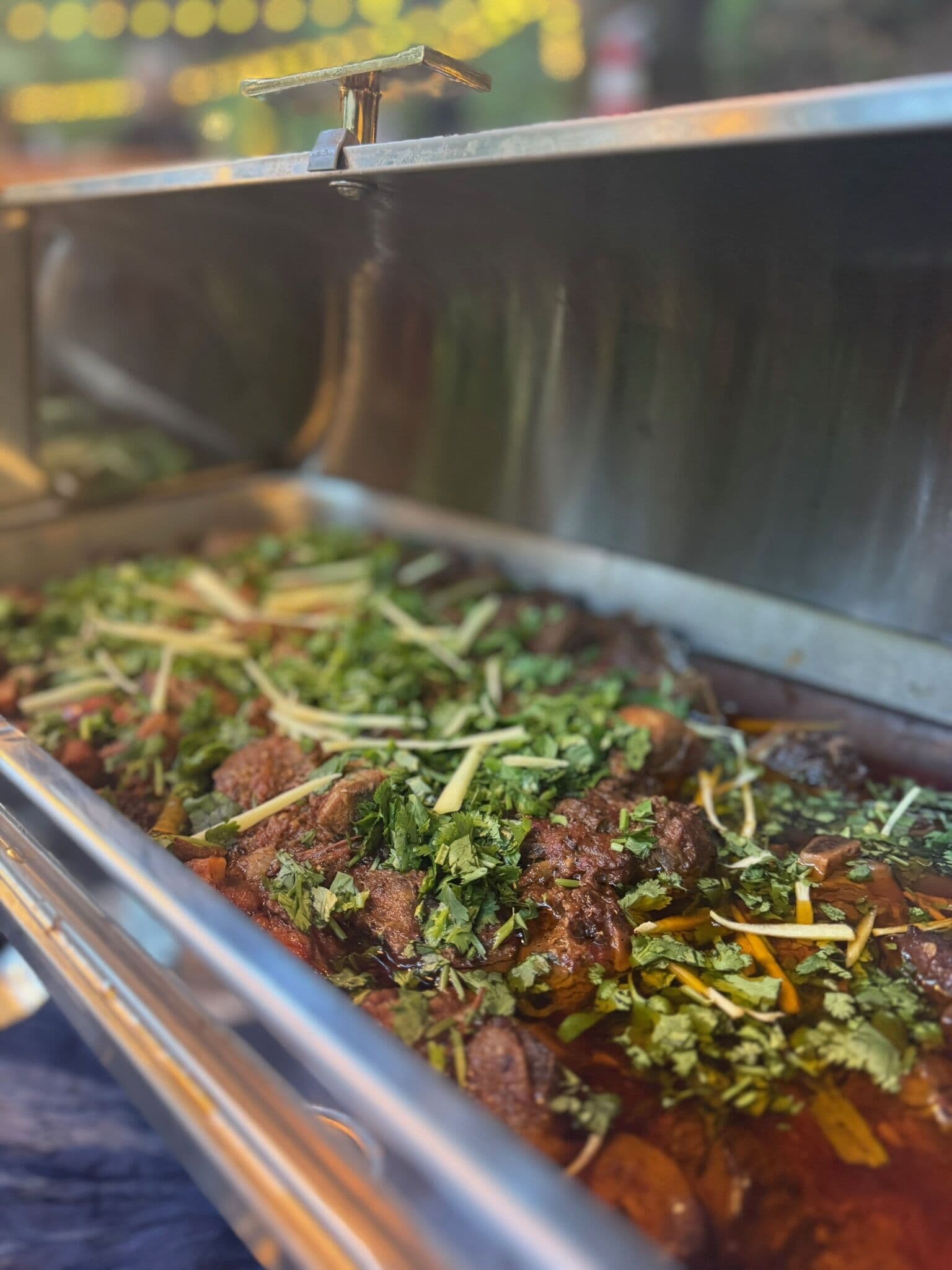 A stainless chafing dish of slow-cooked halal meat curry topped generously with chopped cilantro and julienned ginger, from the Ladle n Wok kitchen.