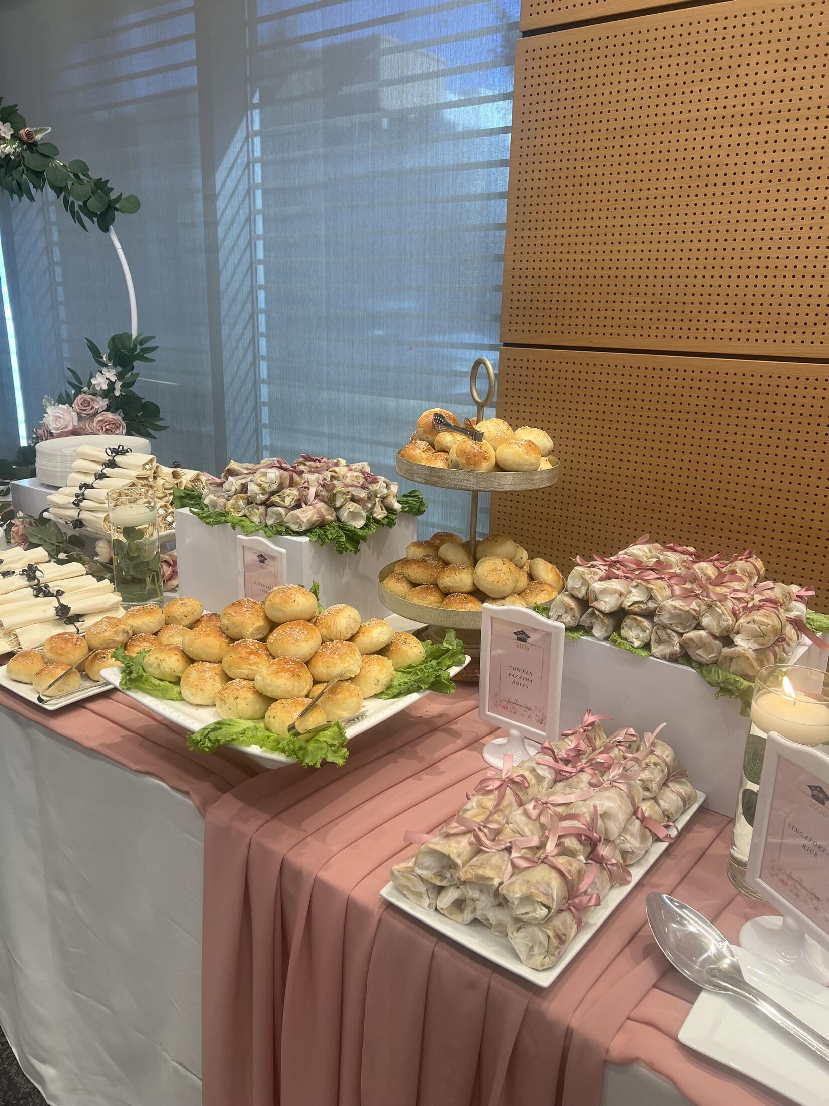 A pastel-styled buffet table with white platters of stuffed rolls, sesame buns, pinwheel wraps and pastries over a blush runner, with a tiered stand and a lit candle at an indoor event.