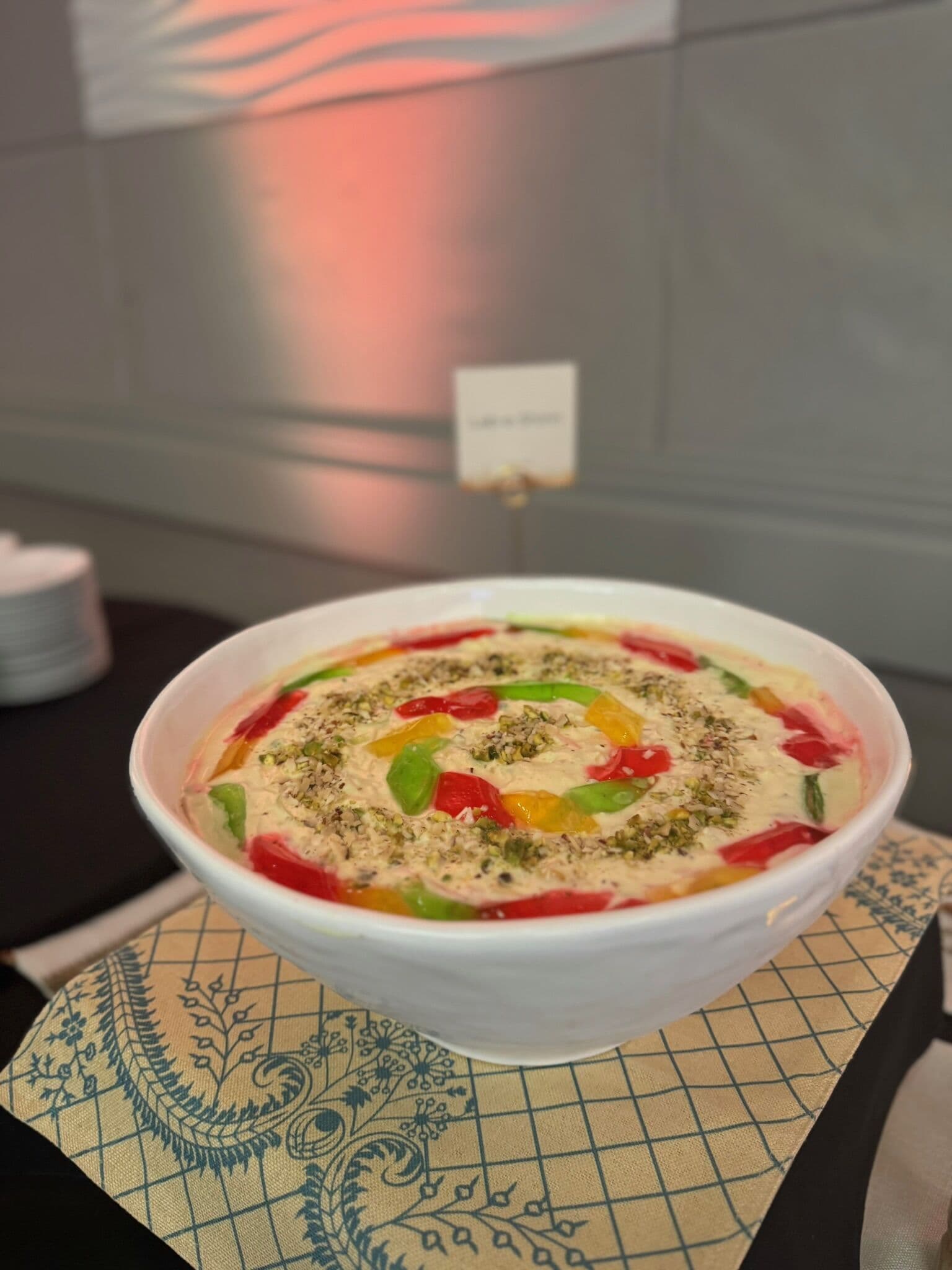 A wide white bowl of creamy set dessert, likely a halal fruit trifle or custard, ringed with red and green glace fruit and dusted with crushed pistachio, resting on a block-printed napkin at a Bay Area event.