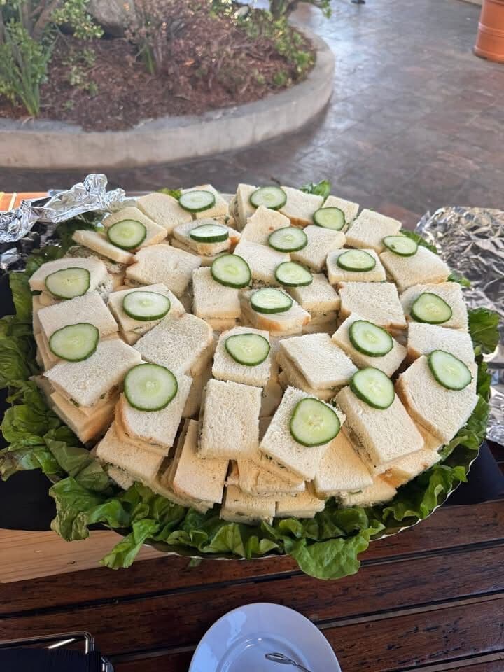 A round platter of crustless finger sandwiches cut into small squares, each topped with a slice of cucumber and laid over a bed of green lettuce, styled for a Hi-Tea spread.