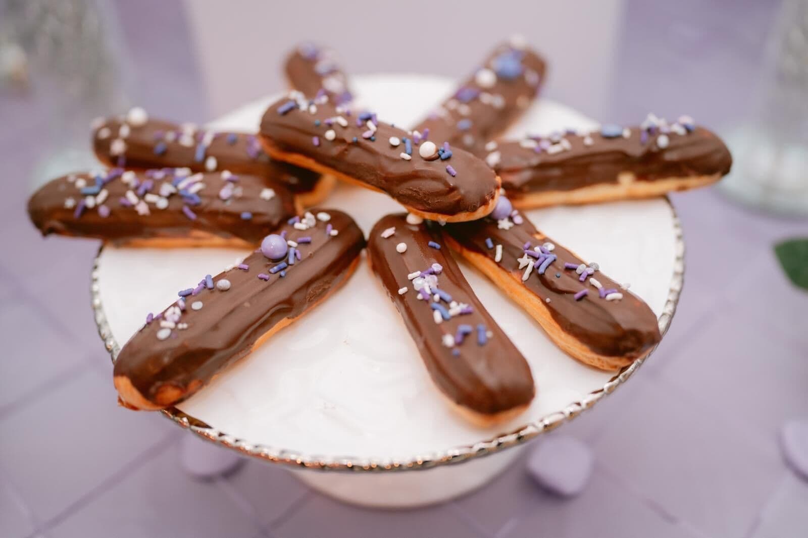 Chocolate-glazed eclairs finished with pastel purple and white sprinkles, fanned in a starburst on a footed white cake stand at a dessert table.