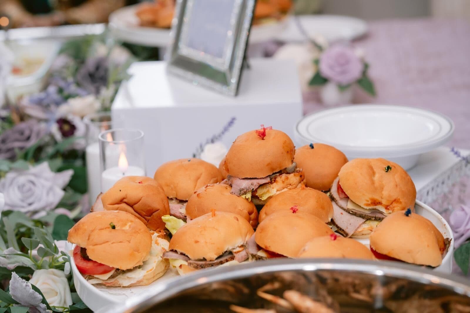 A platter of soft dinner-roll sliders layered with sliced deli meats, cheese and tomato, garnished with confetti picks and set among lavender florals and a votive candle at a Bay Area event.