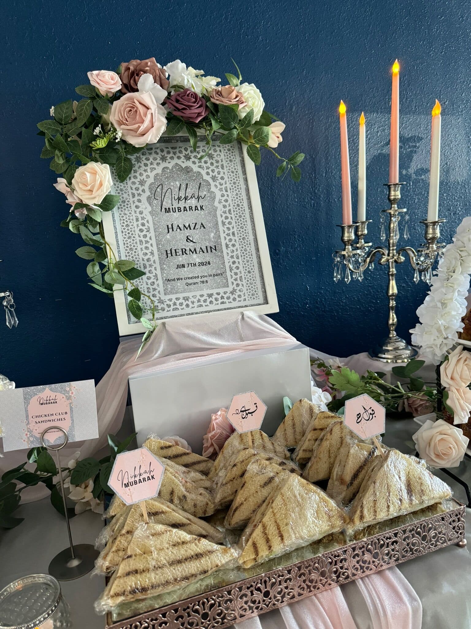 A footed tray of grilled halal sandwich triangles pressed with panini marks, styled under a floral arch with a Nikkah Mubarak sign and lit taper candles.