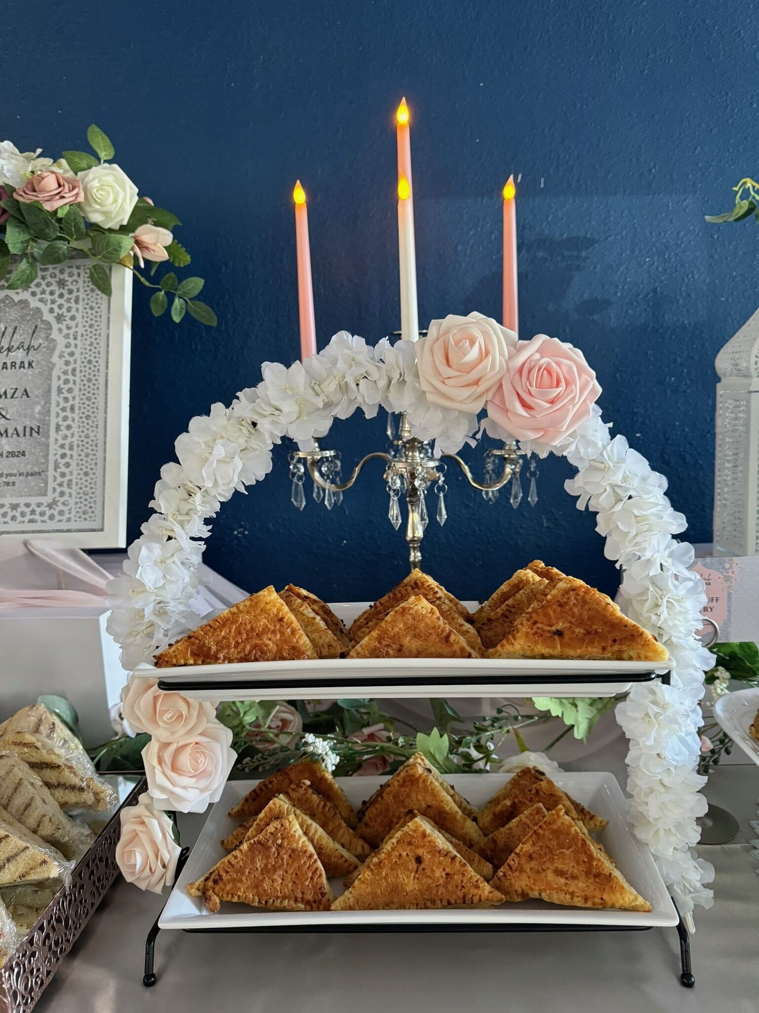 Two tiered white trays of golden grilled sandwich triangles set beneath a floral-wrapped candelabra with pink taper candles against a navy backdrop.