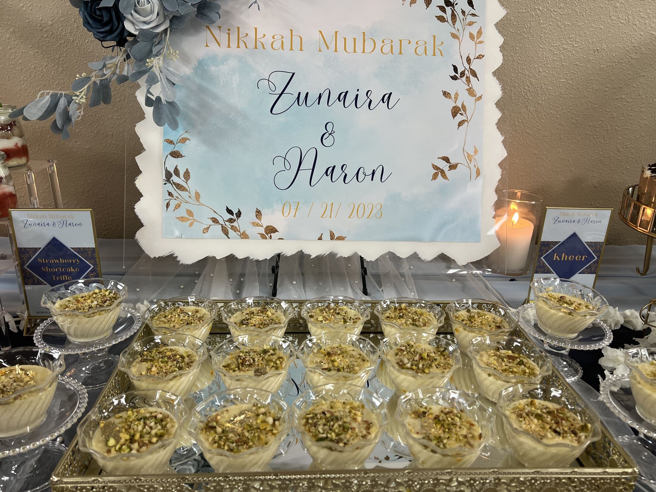 A tray of individual kheer cups topped with chopped pistachio, set on a mirrored platter beneath a Nikkah Mubarak sign for Zunaira and Aaron alongside labeled dessert station cards.