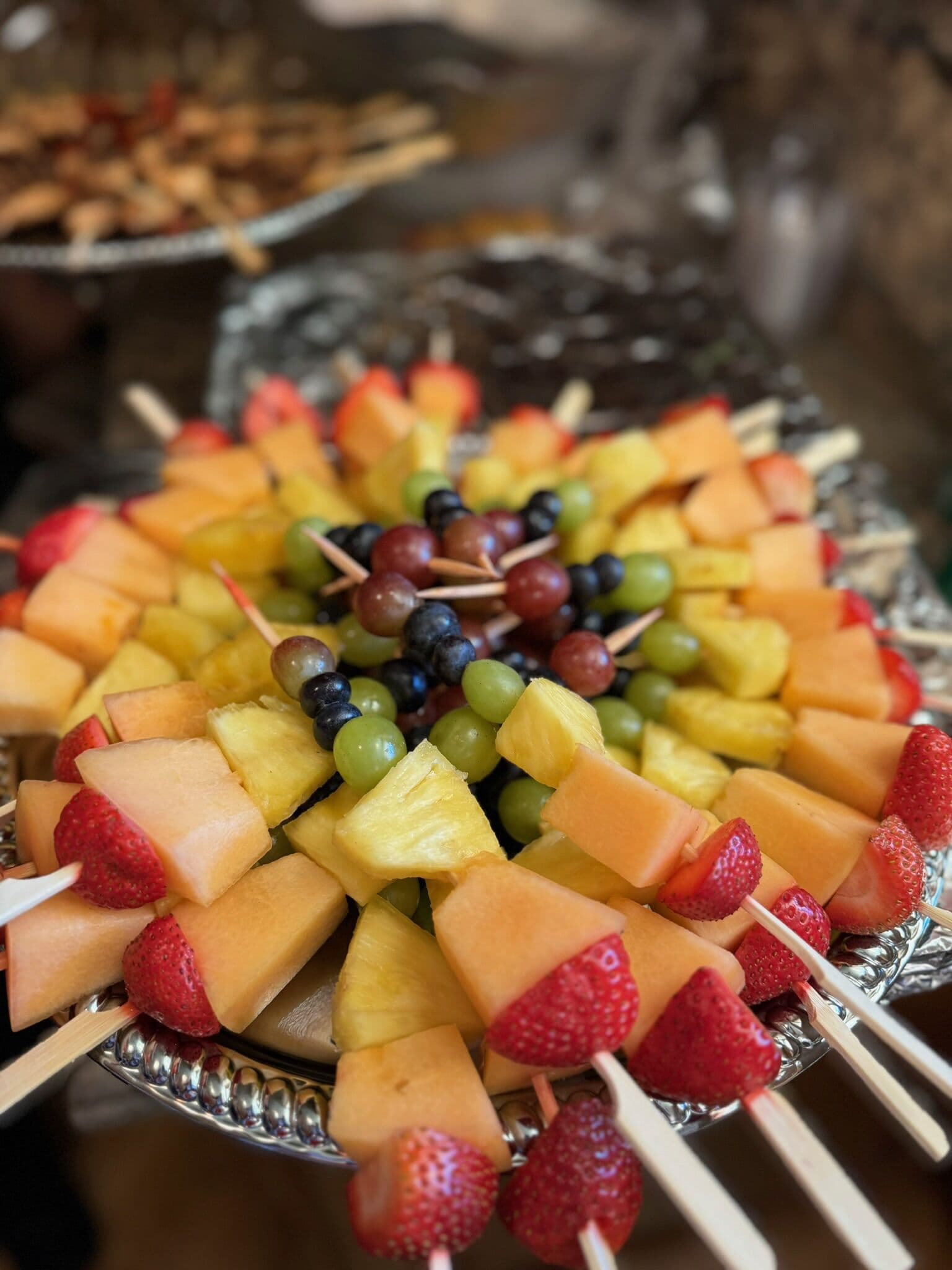 A silver platter arranged with skewers of fresh fruit, including strawberries, cantaloupe, pineapple, and green and dark grapes, radiating from the center at a catered spread.