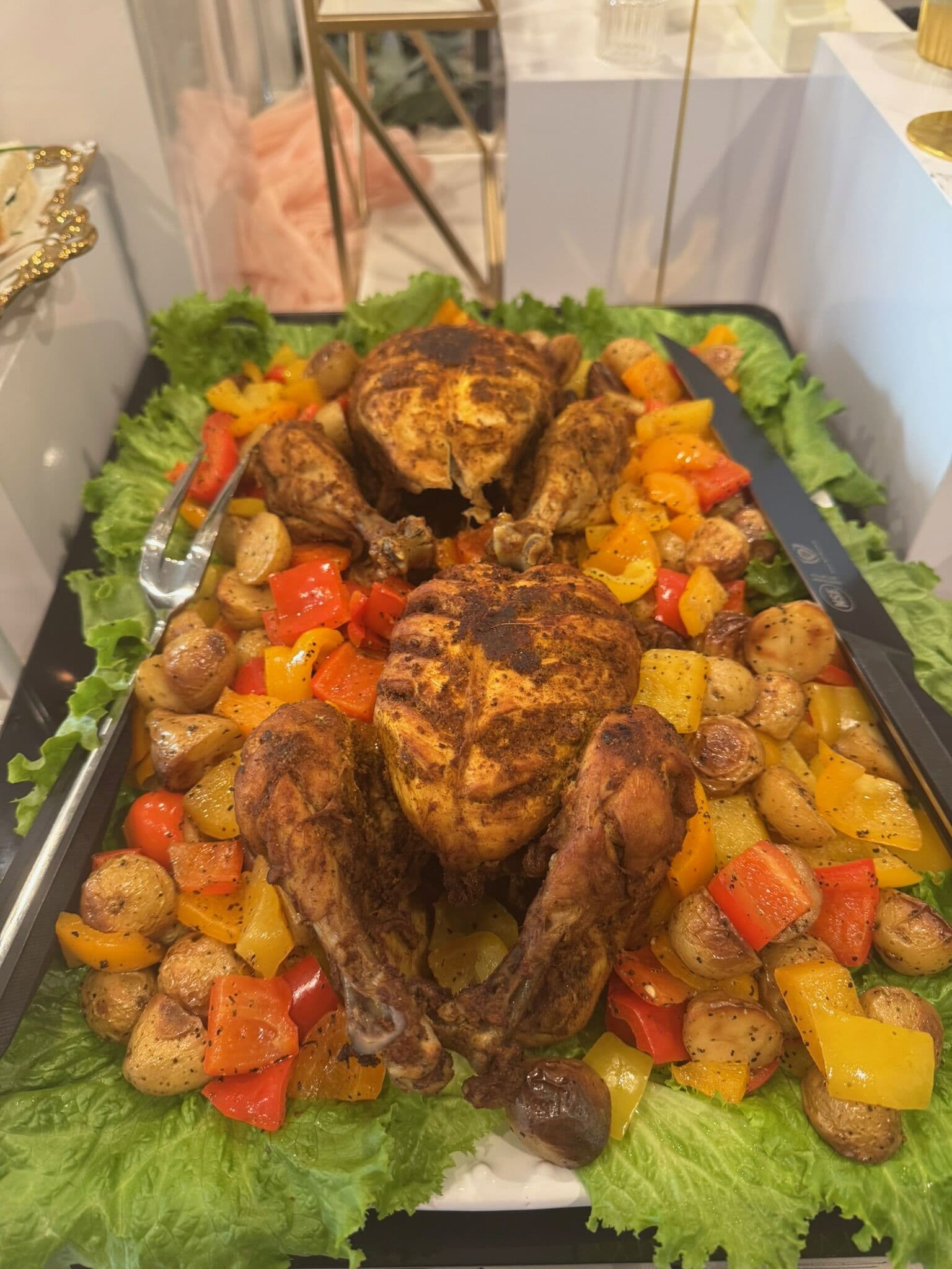 A platter of whole roast halal chargha chickens rubbed with spice, plated over lettuce with roasted baby potatoes and red and yellow bell peppers.