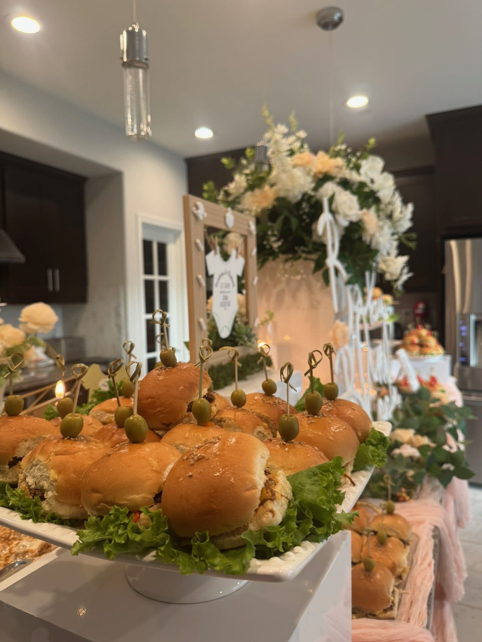 A white pedestal platter of glazed slider buns filled with meat and lettuce, each pinned with a skewered green olive, set against a floral arch and mirror backdrop at a Bay Area shower.