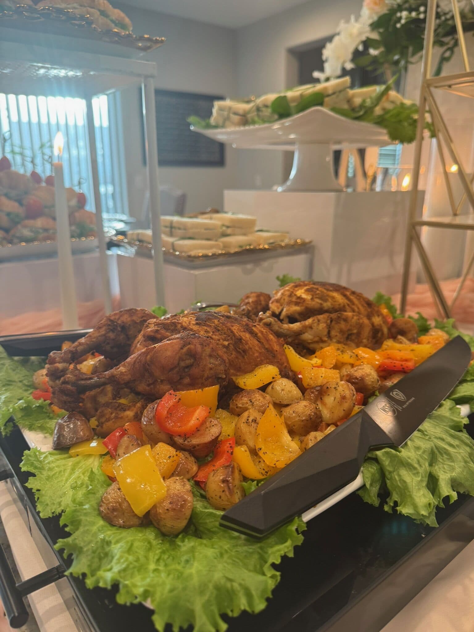A tray of whole spice-rubbed roast halal chickens plated over lettuce with roasted potatoes and bell peppers, a carving knife alongside, with tea sandwiches tiered behind.
