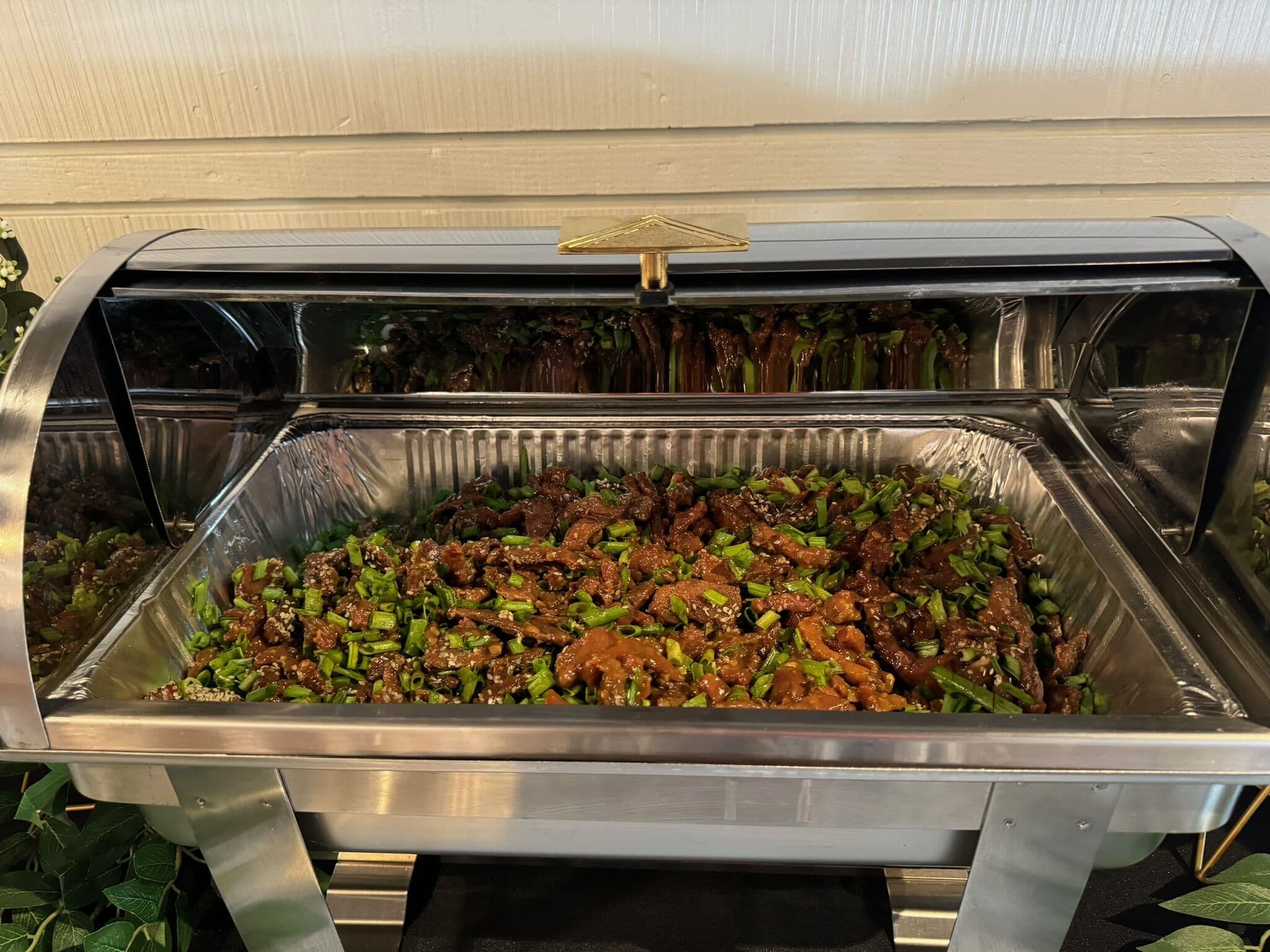 A roll-top chafing dish holding halal beef in a dark sauce scattered with sesame and sliced scallions on a catering buffet.