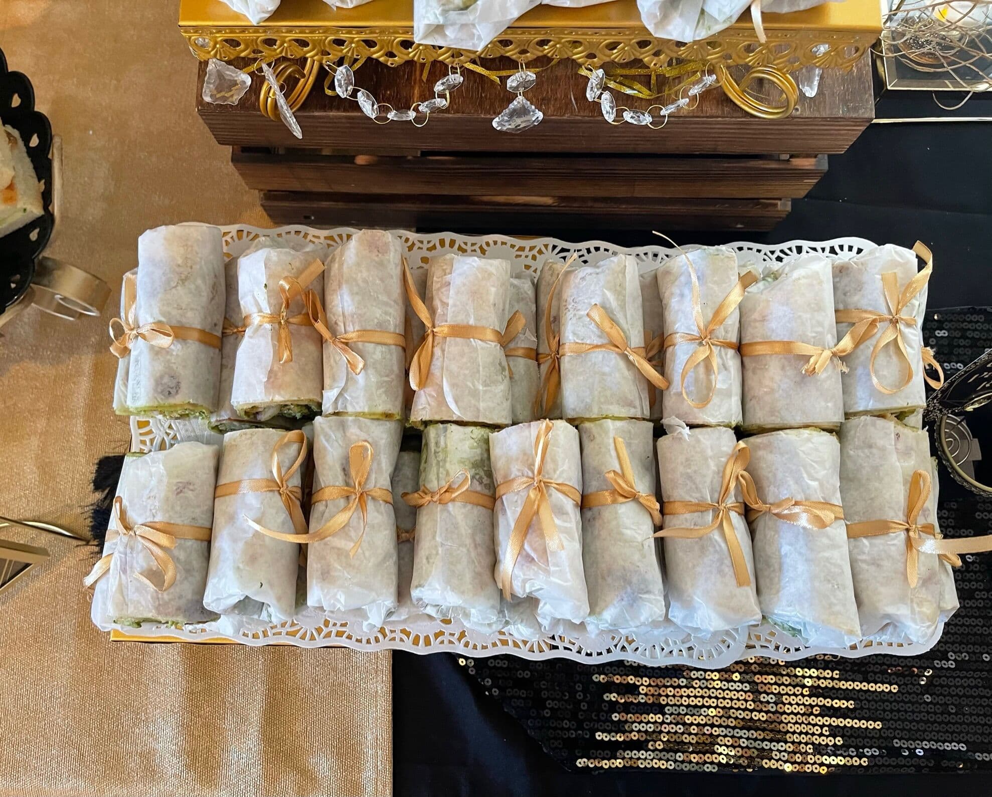 Rows of wrap rolls in parchment paper, each tied with a gold ribbon and lined up on a doily-lined tray over a black sequined cloth.