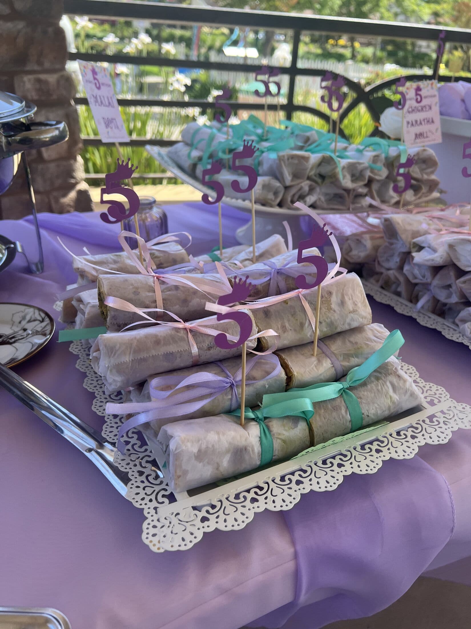 Stacked paratha rolls wrapped in paper and tagged with pastel ribbon and purple crown picks, priced on a doily-lined tray at an outdoor lilac-themed party.